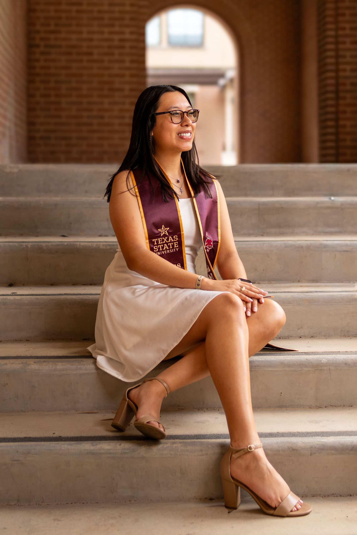 A smiling woman in a Texas State graduation stole sits on stone steps in a beige dress and heeled sandals.