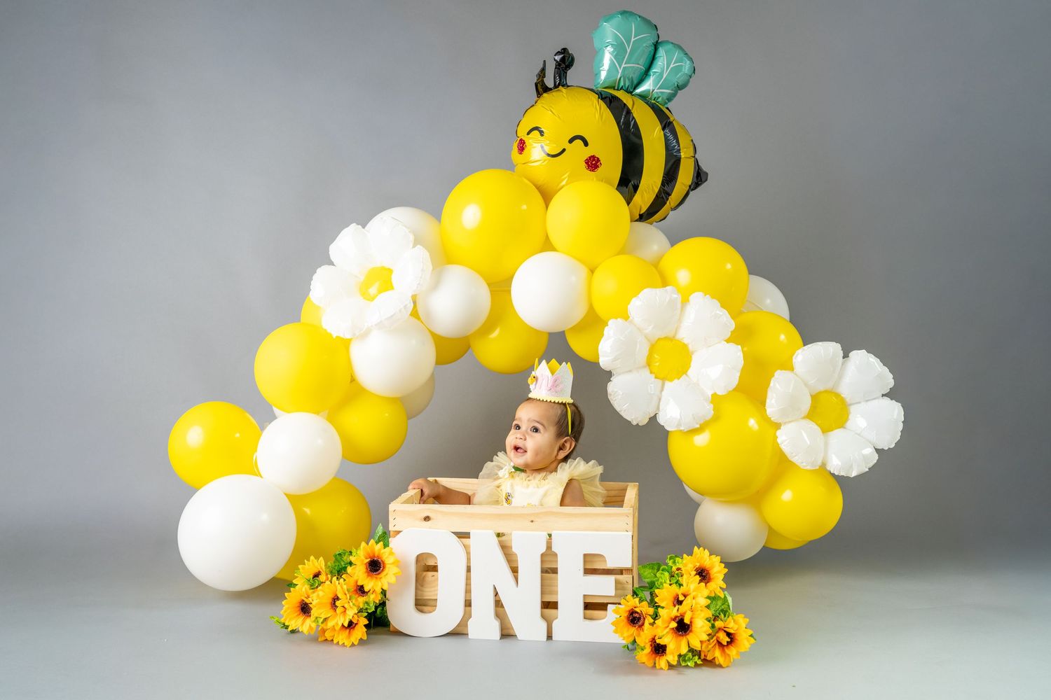 Baby's first birthday cake smash with yellow and white balloon arch, bee balloon, daisy balloons, and ONE sign with sunflowers.