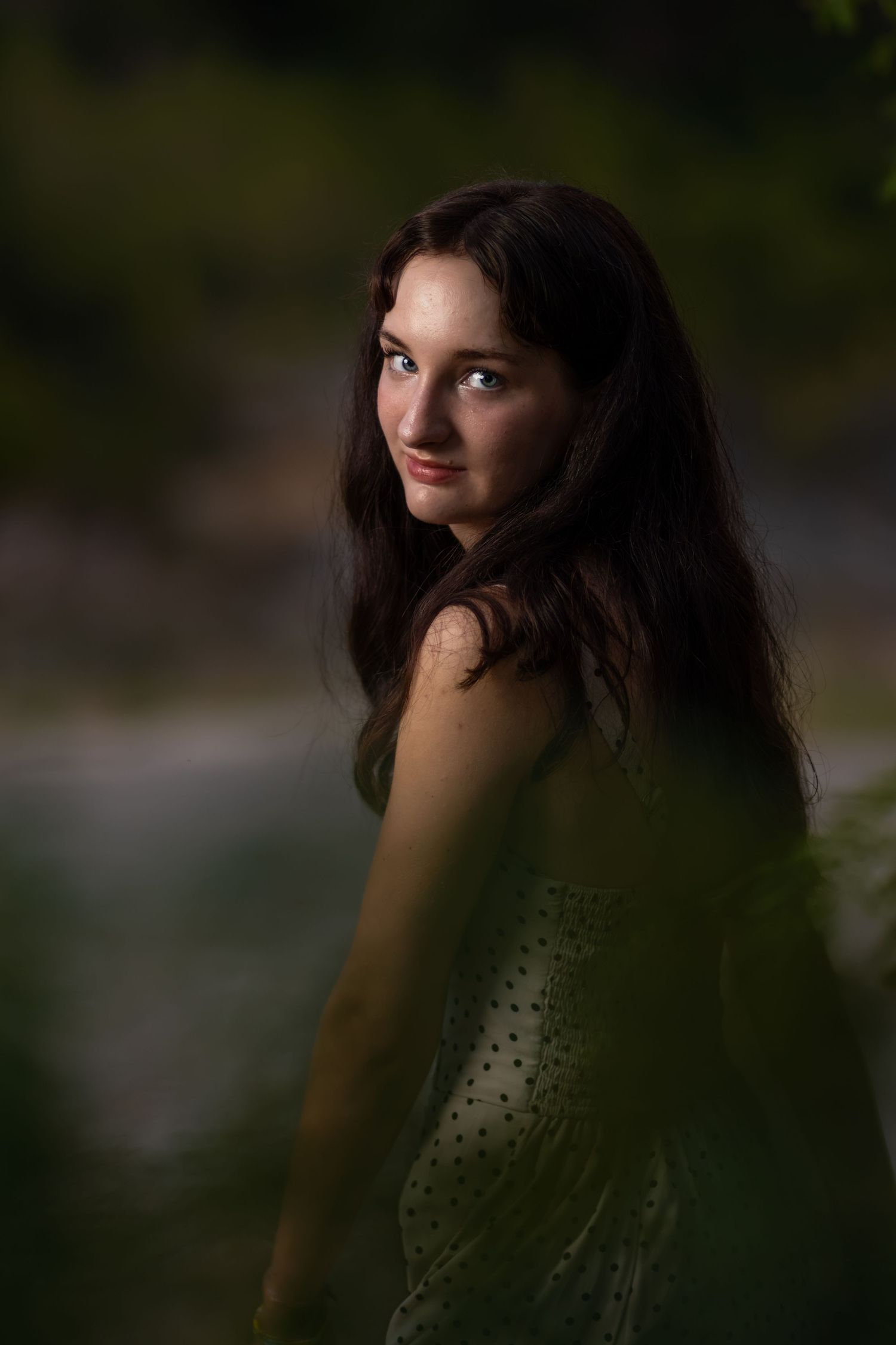 Young woman with long dark hair and blue eyes in a green dress, posing outdoors with a blurred natural background.