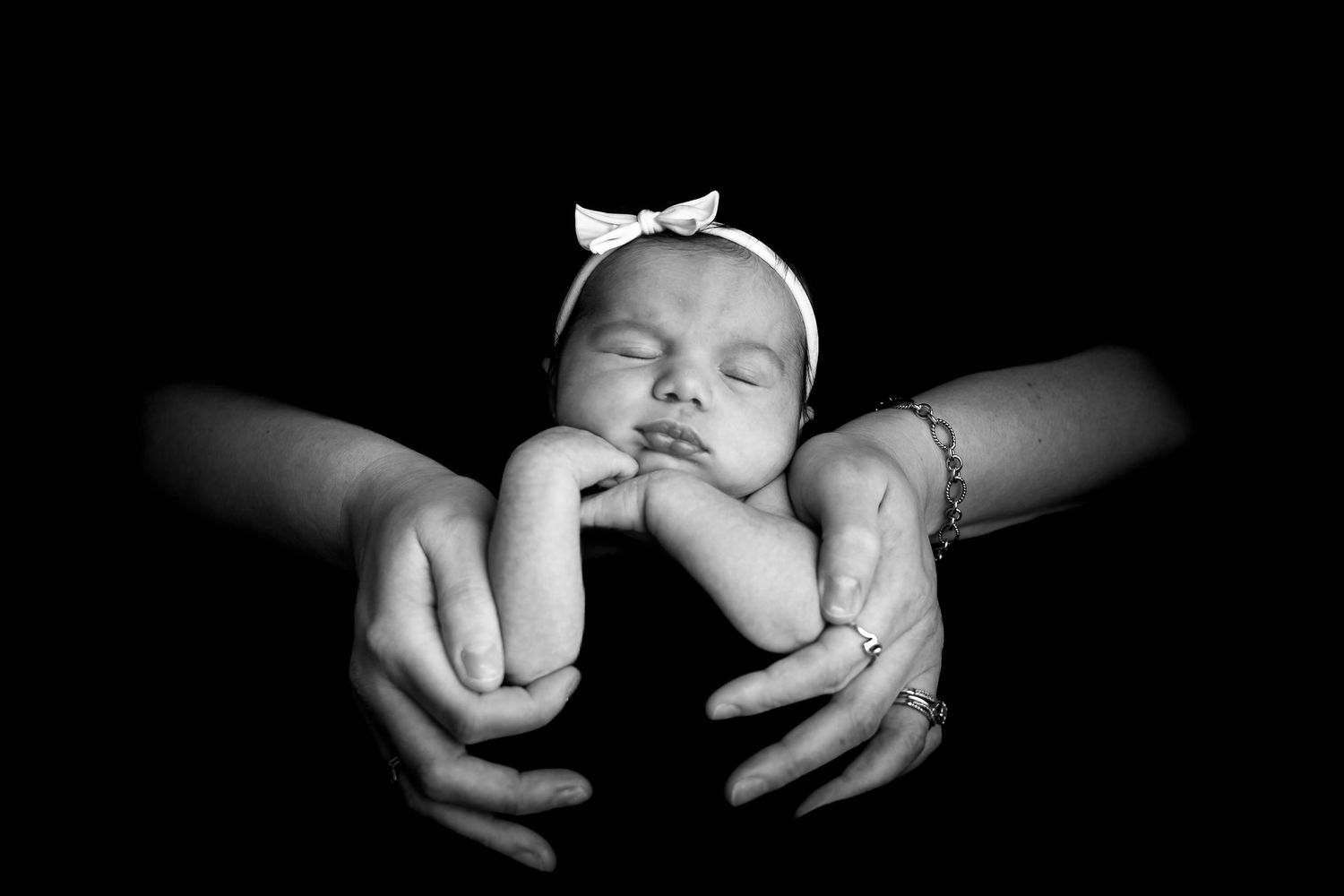 Black and white photo of a sleeping newborn baby with bow headband cradled in parents hands.