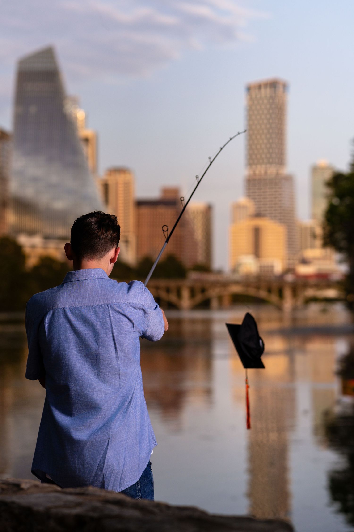 Man fishing with a rod by a river with a blurred city skyline in the background at sunset.