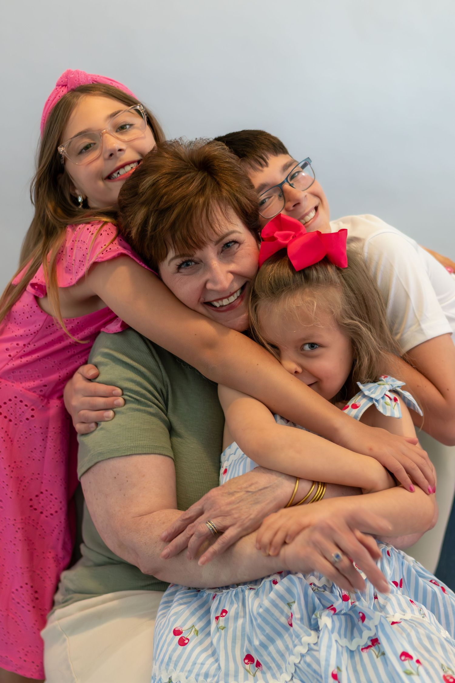 A grandmother smiles warmly while three young children hug her tightly in a joyful family portrait.
