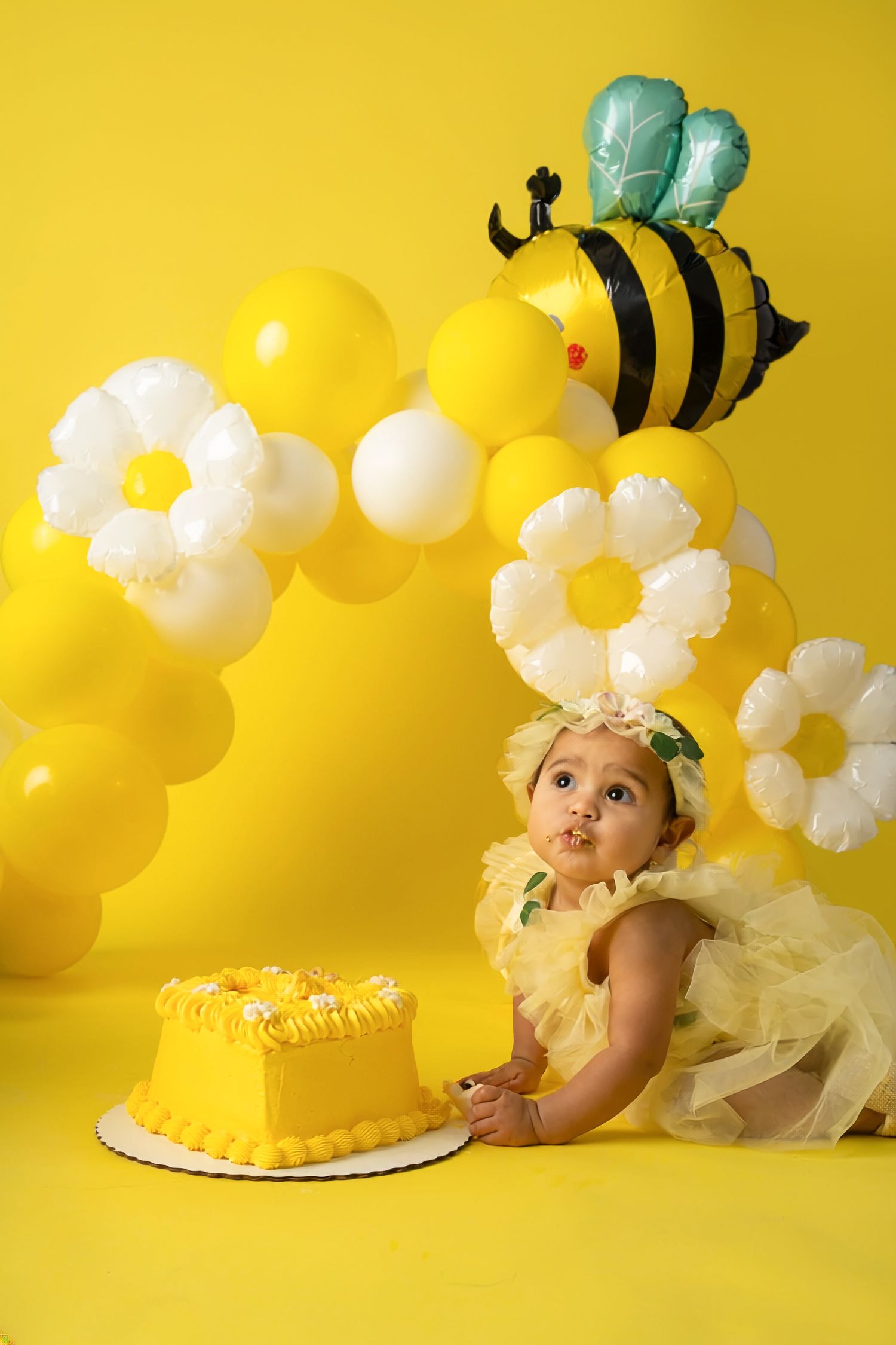 Baby girl in yellow tutu celebrates first birthday with yellow cake, bee balloon, and daisy balloon decorations.