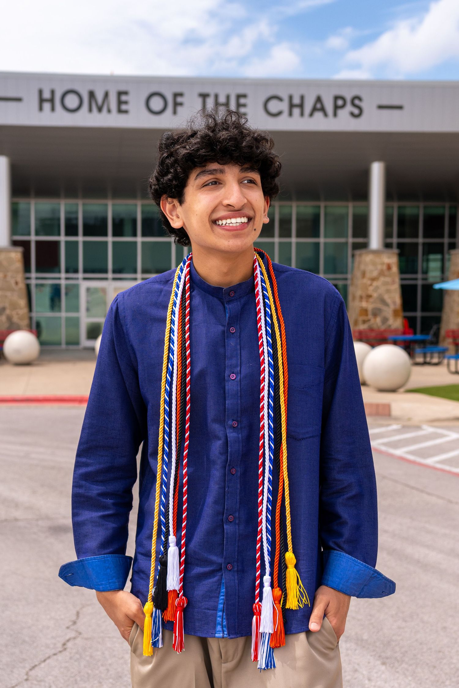 Smiling graduate wearing colorful honor cords stands in front of Home of the Chaps school building on a sunny day.