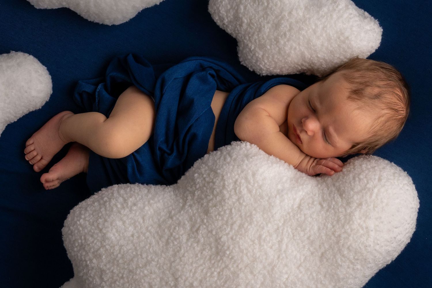 Newborn baby sleeping peacefully on blue fabric surrounded by fluffy white cloud props in a dreamy studio photo.