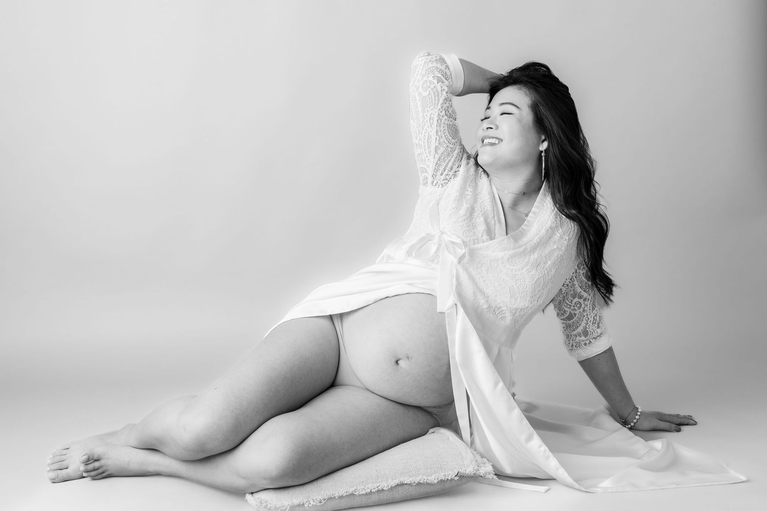 Black and white maternity photo of a woman in a white lace robe sitting gracefully on the floor.