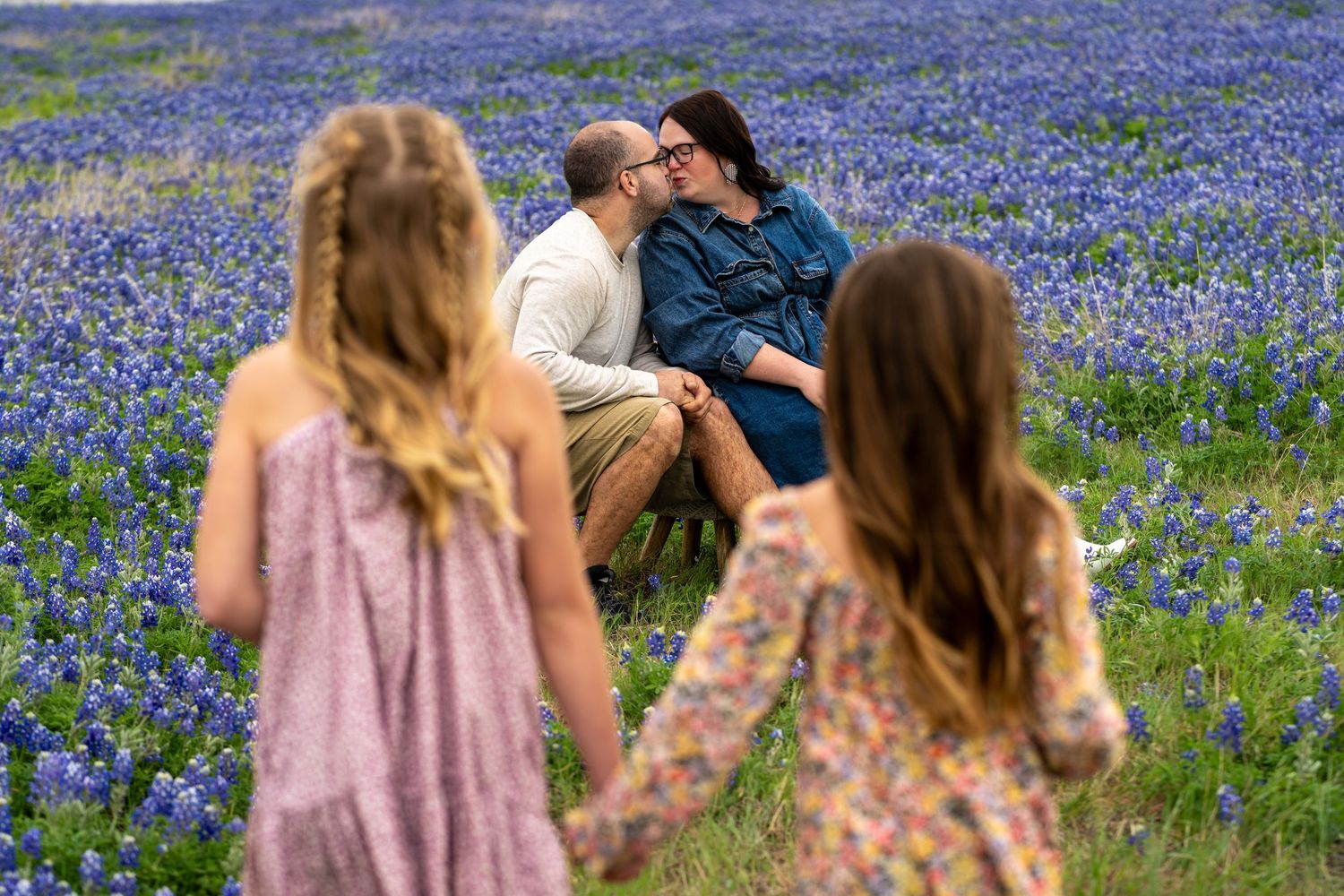 Couple kissing in a field of Texas bluebonnets while two girls in floral dresses watch in the foreground.