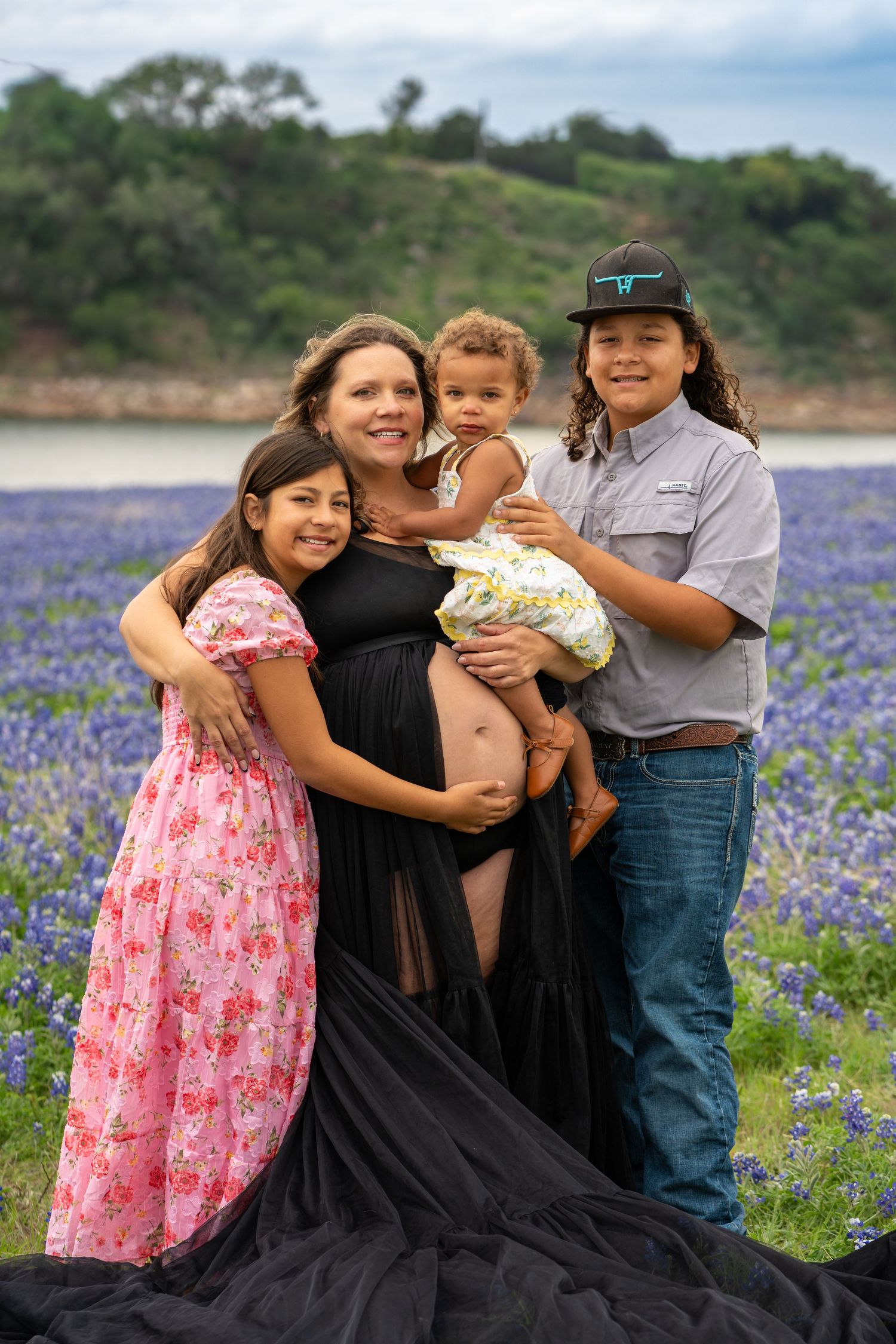 Pregnant woman poses with three children in a bluebonnet field in Texas during a maternity photo session.