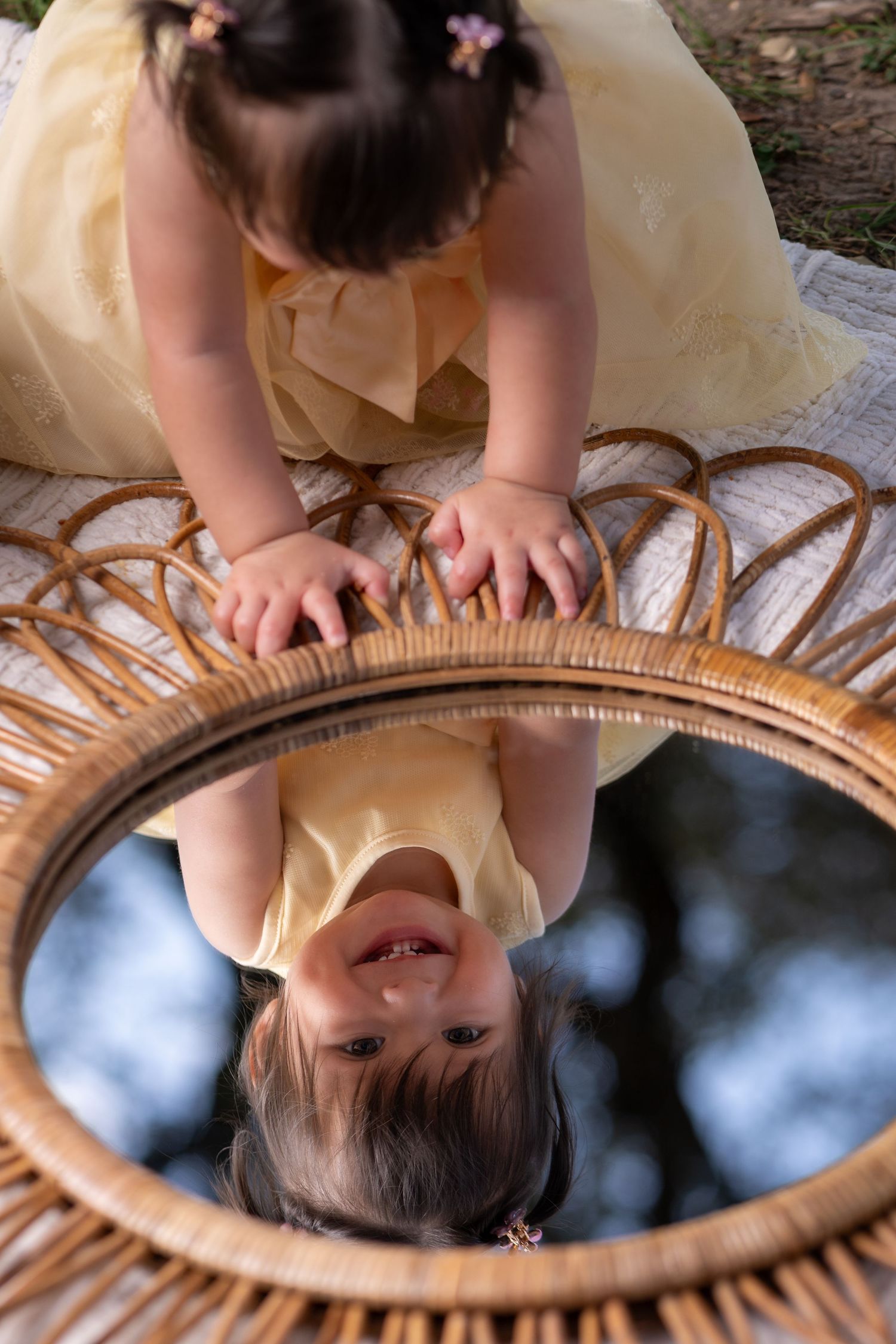 A toddler girl in a yellow dress looks at her reflection in a round rattan mirror outdoors.