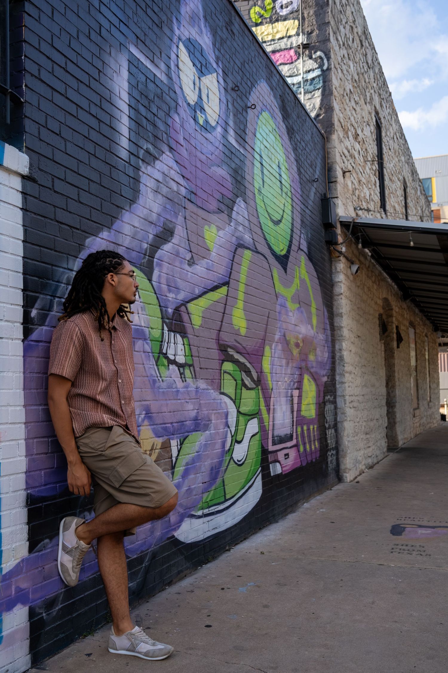 Young man with long braids leans against a colorful graffiti mural wall in an urban alley wearing shorts and sneakers.