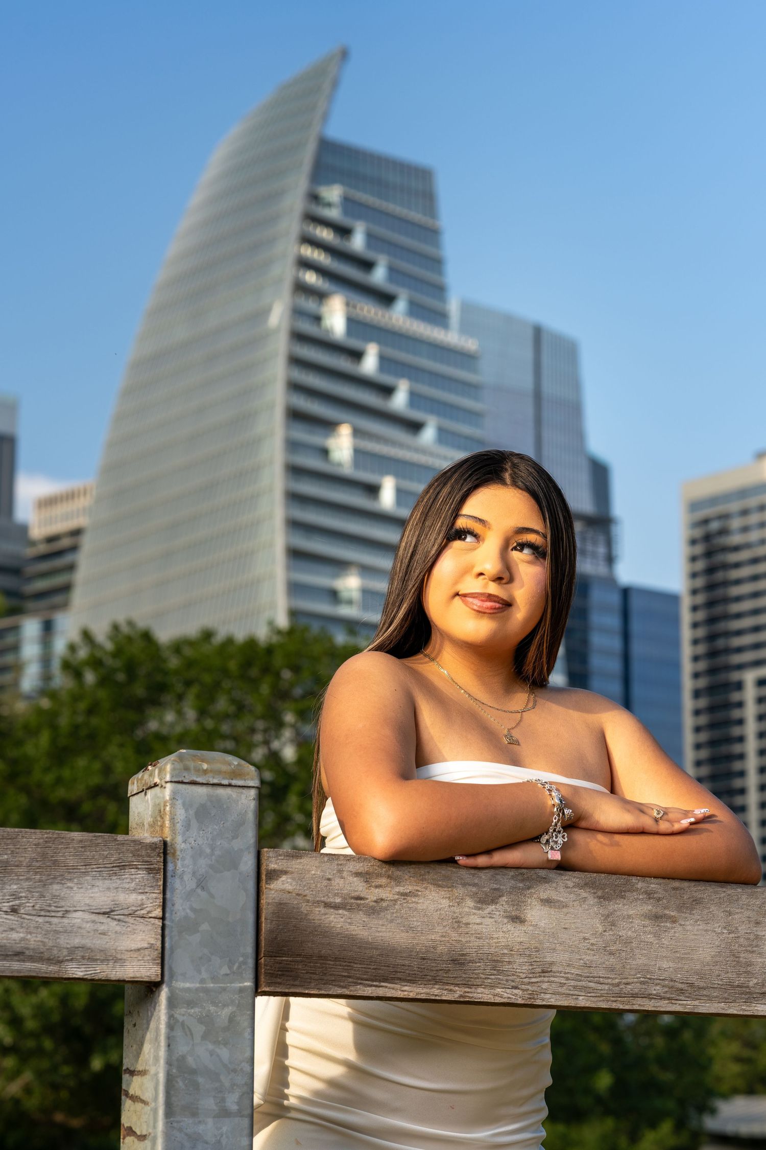 Young woman in white dress leans on wooden fence with modern city skyscrapers in background on sunny day.