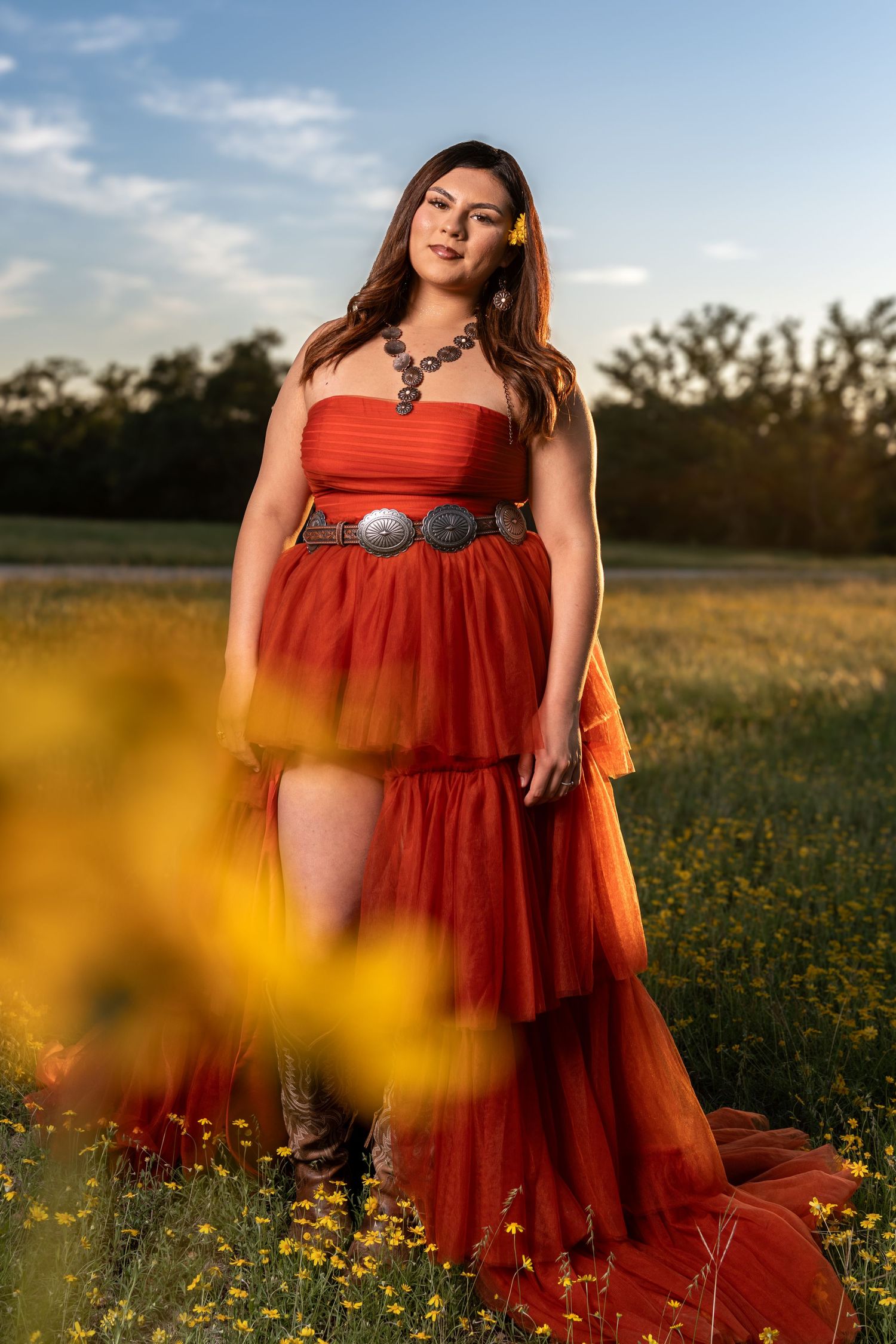 Woman in rust orange tulle gown with concho belt standing in a sunny wildflower field.