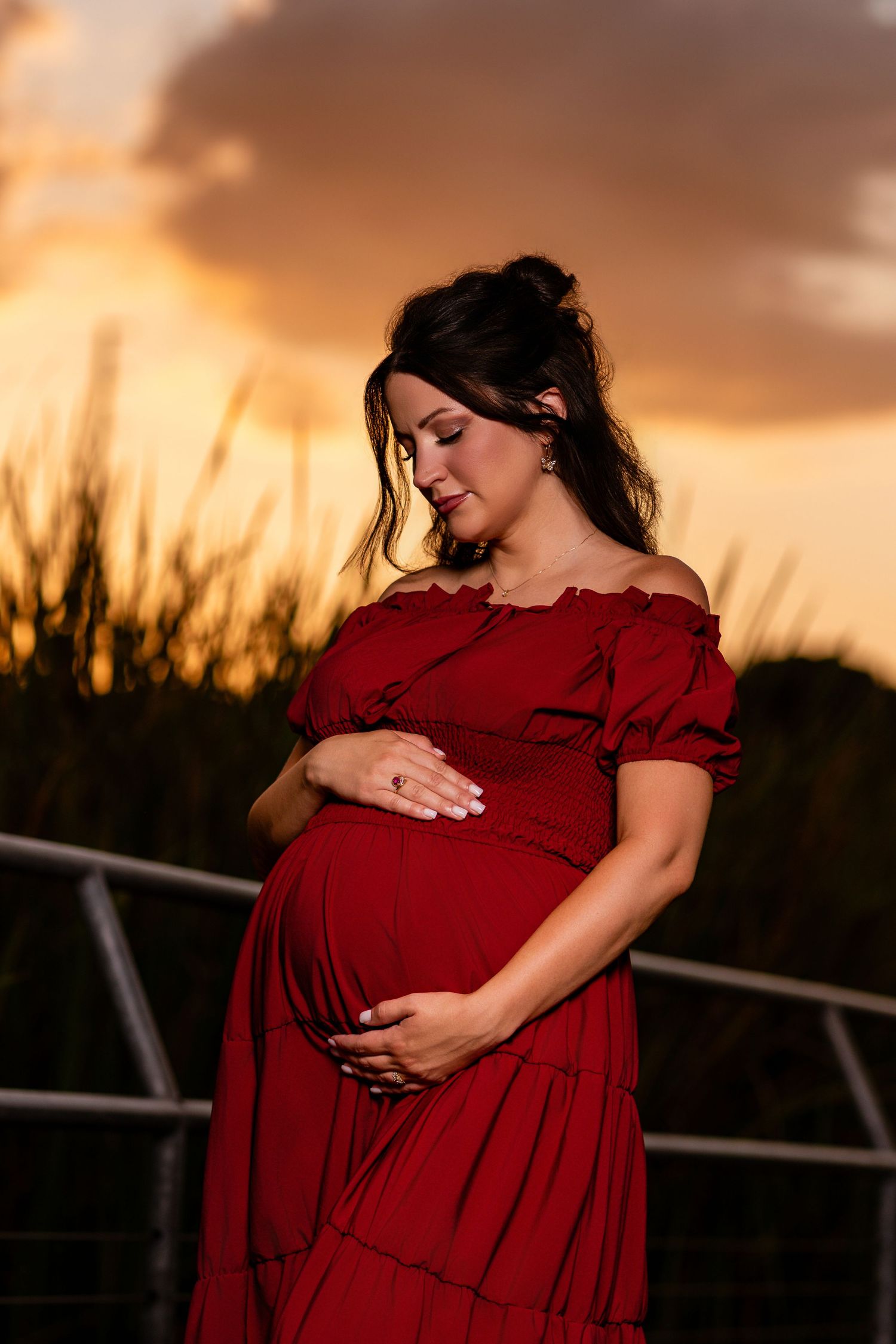 Pregnant woman in red off-shoulder dress cradling her baby bump at golden sunset outdoors.