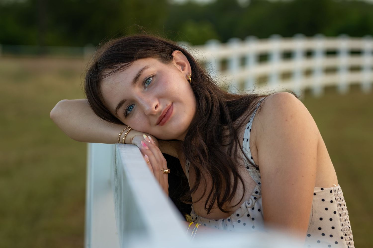 Young woman with brown hair leans on white fence, smiling softly outdoors in natural light.