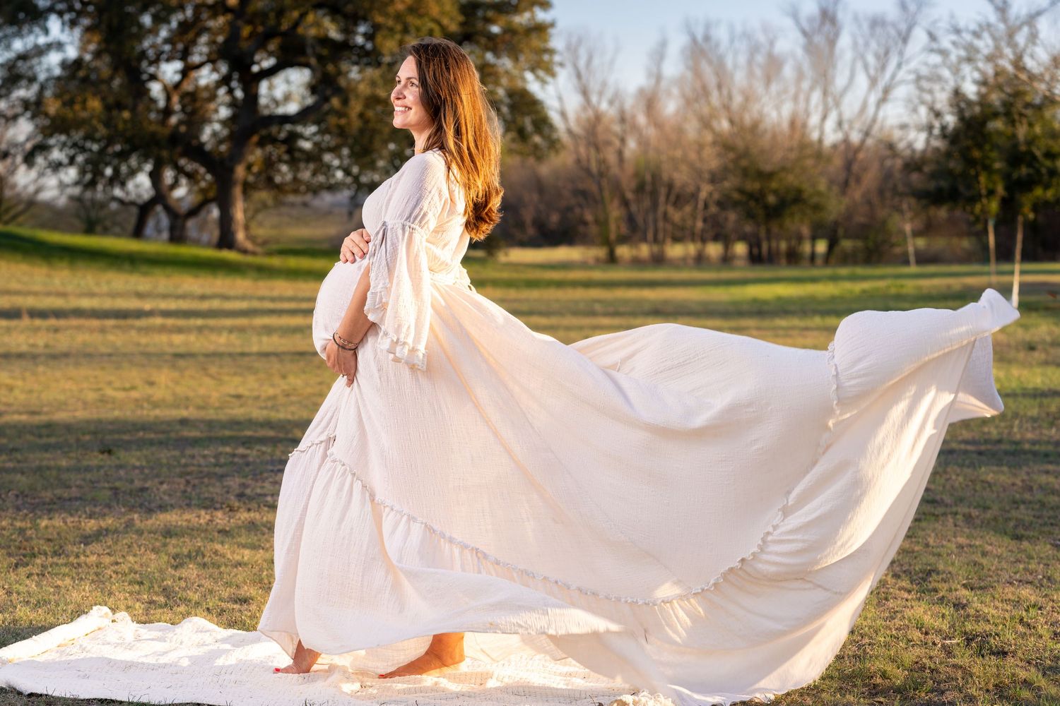 A flowing white maternity dress billows in the breeze as someone poses barefoot in a sunlit park at golden hour.