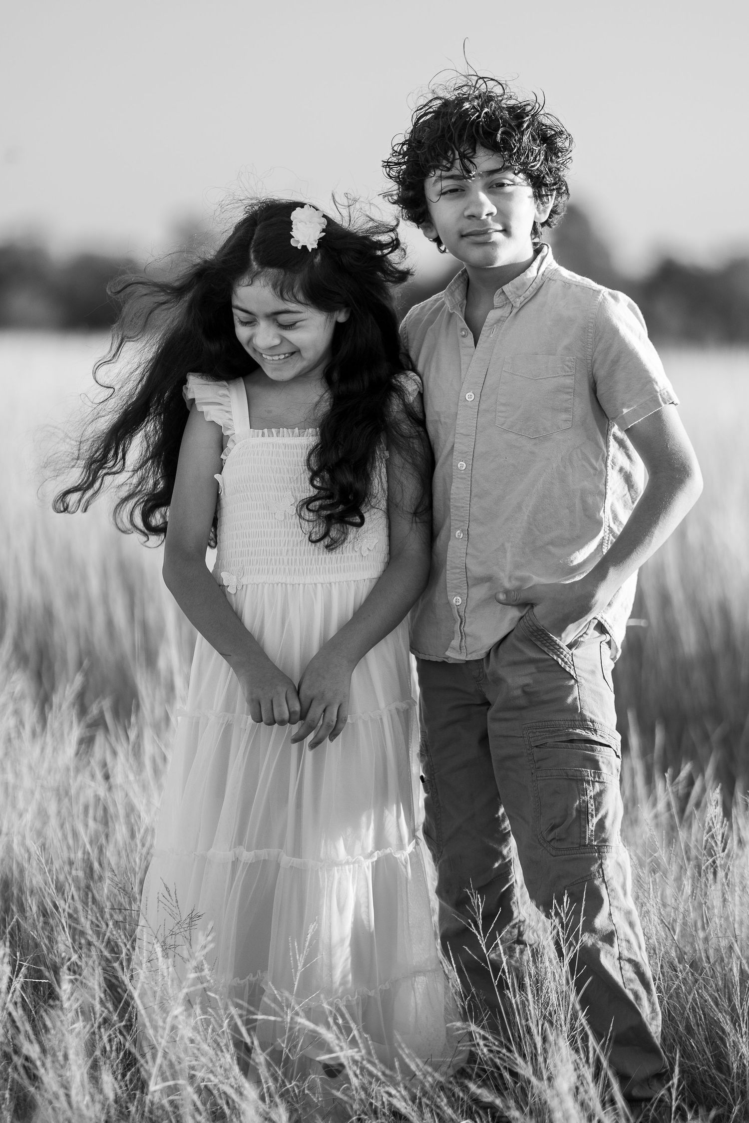 Two children stand together in a sunlit field wearing light colored clothing while wind blows through their hair.