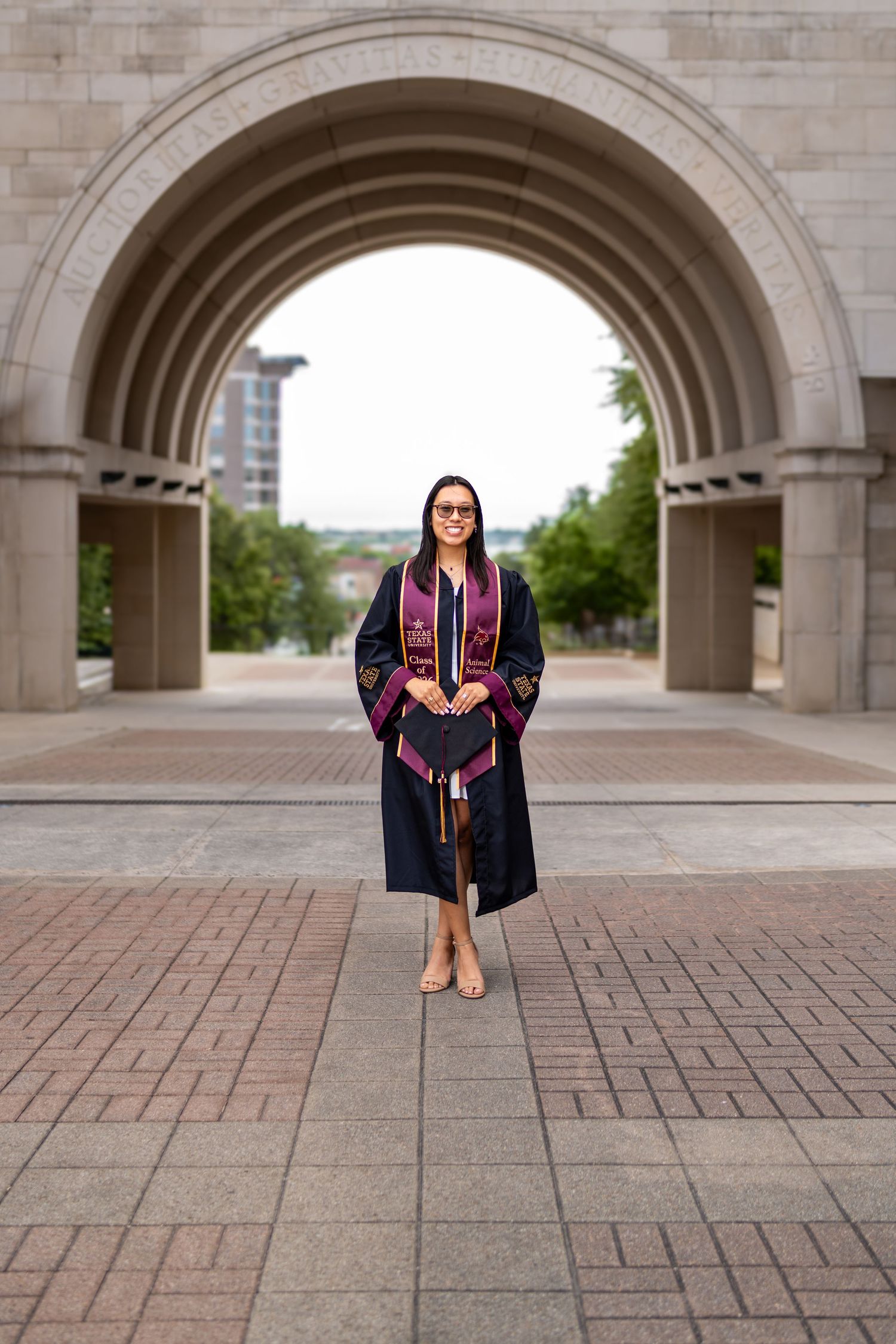 Graduate in black gown and maroon stole stands under stone arch at university campus ceremony.
