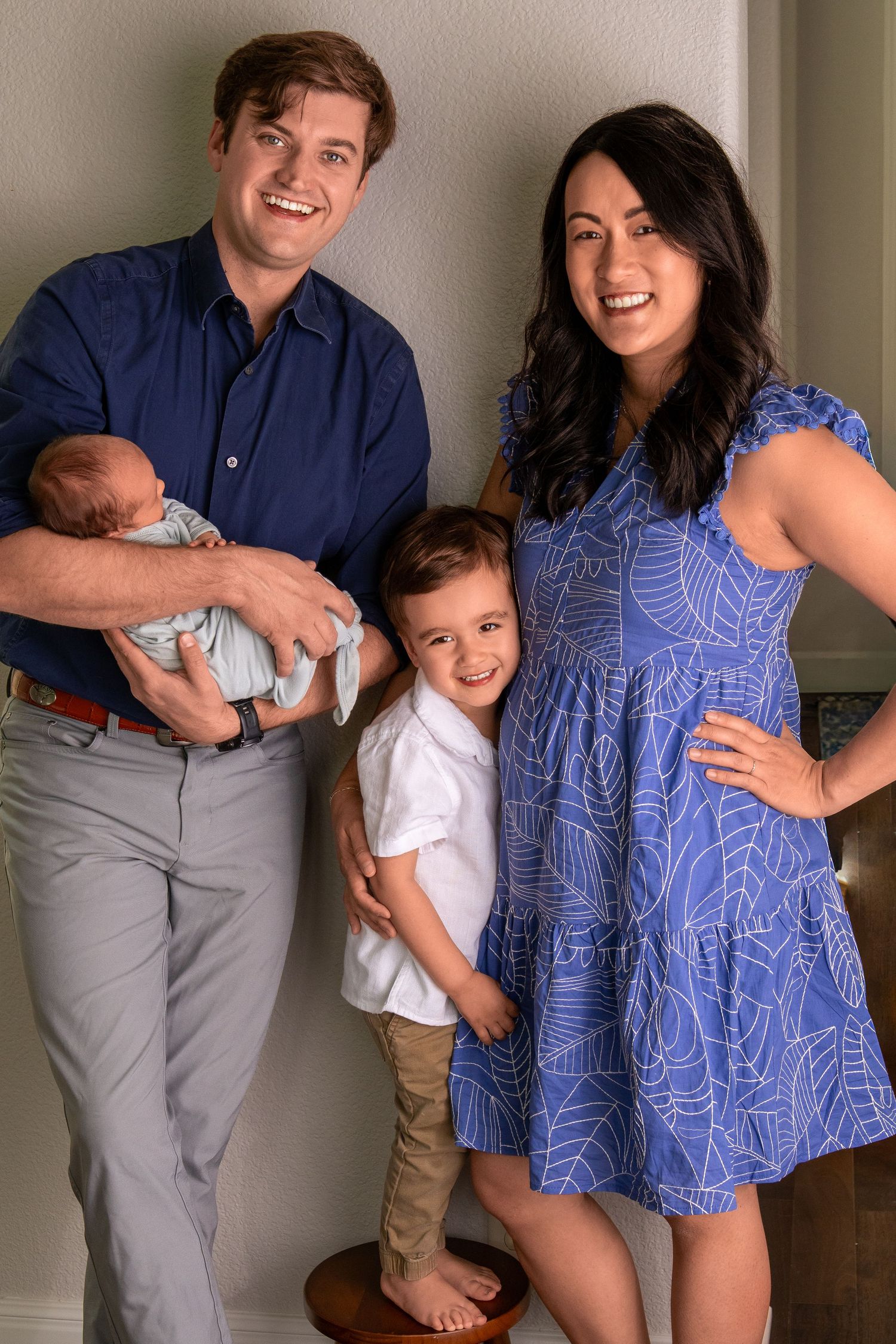 Smiling family of four poses together, dad holds newborn baby, toddler stands between parents.