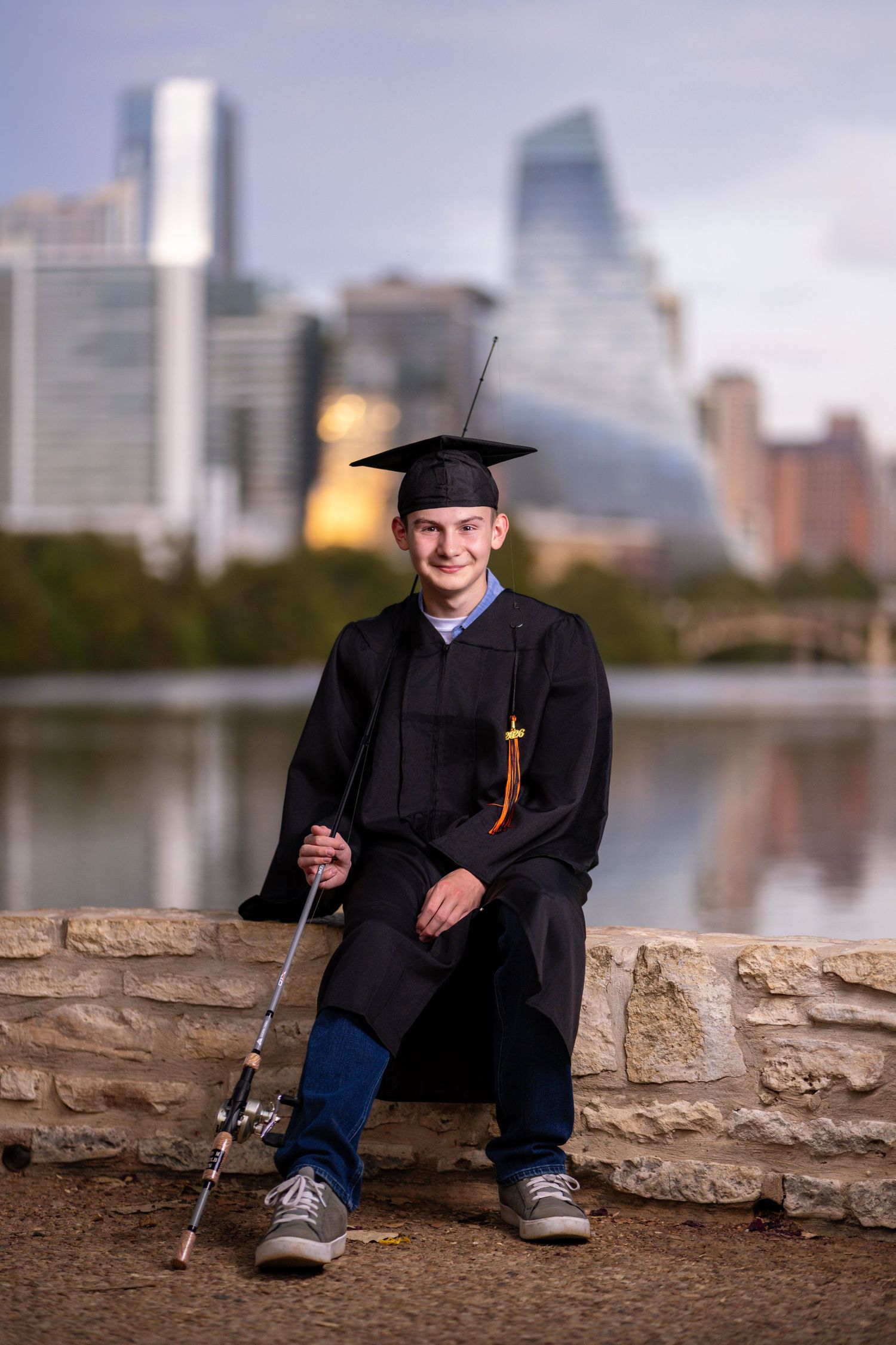 Young man in graduation cap and gown holding a fishing rod, seated by a river with a city skyline backdrop.