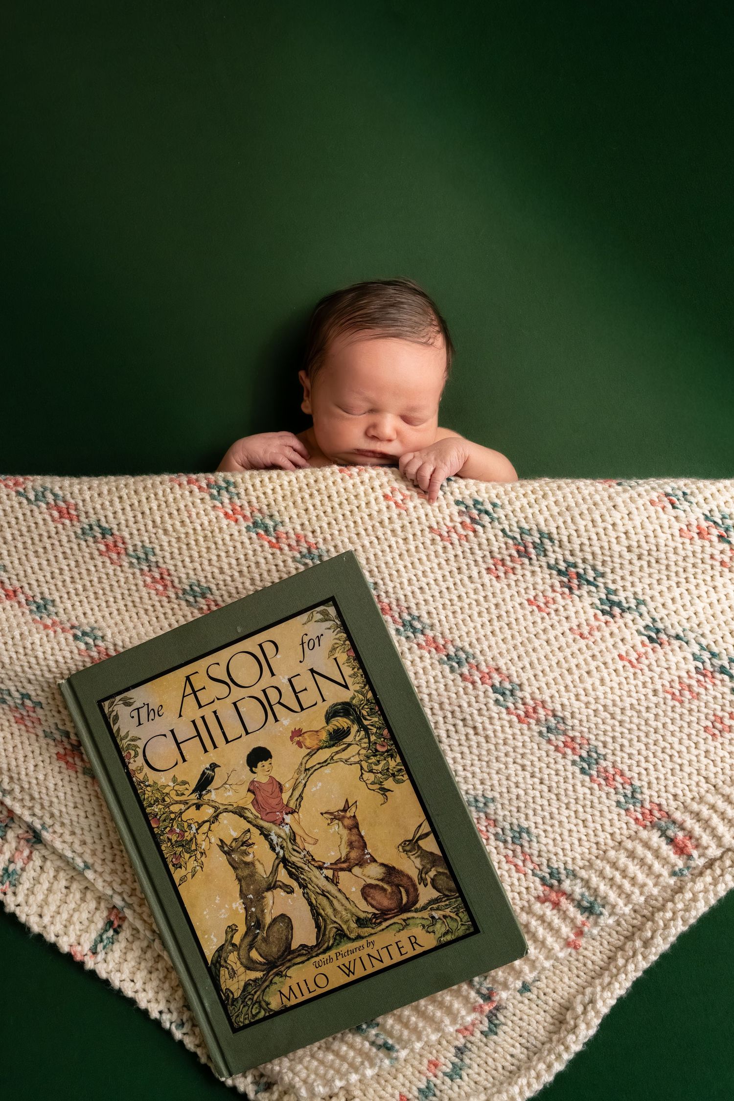 Newborn baby sleeping on a knitted blanket beside a copy of Aesop for Children by Milo Winter.