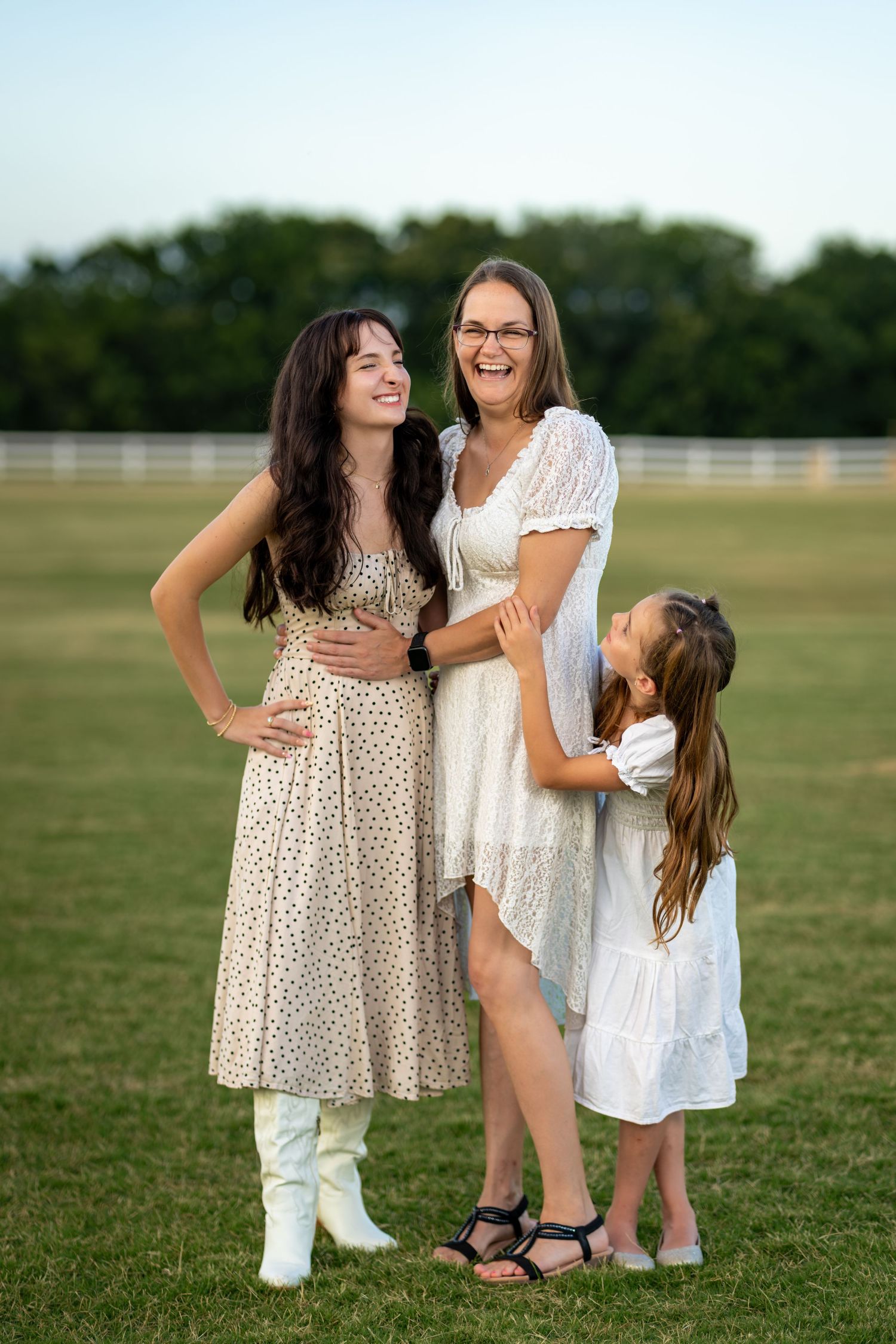 Three females in white dresses laugh together outdoors on green grass near a white fence on a sunny day.
