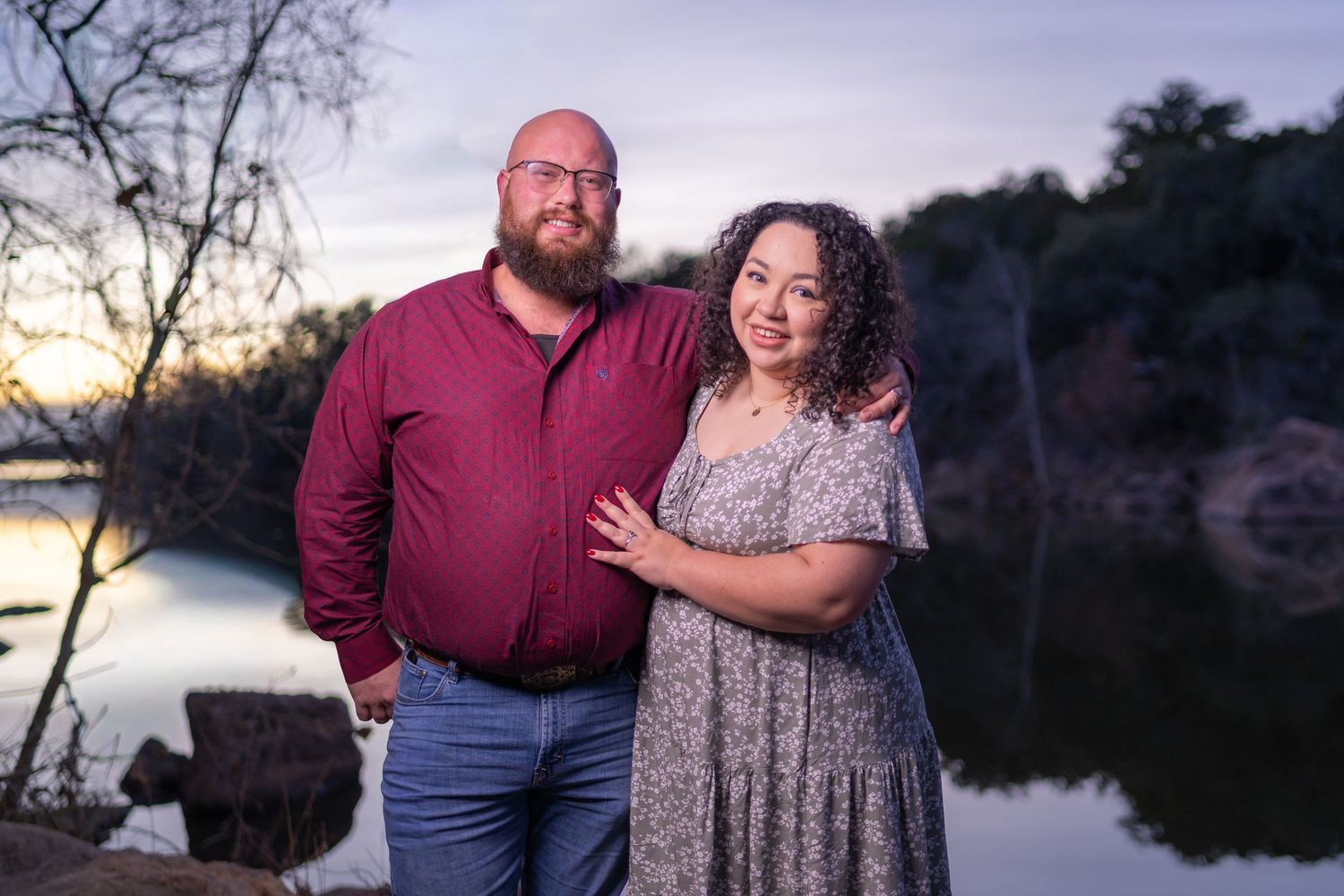 Couple poses together during sunset engagement photoshoot by serene lake with trees in background.