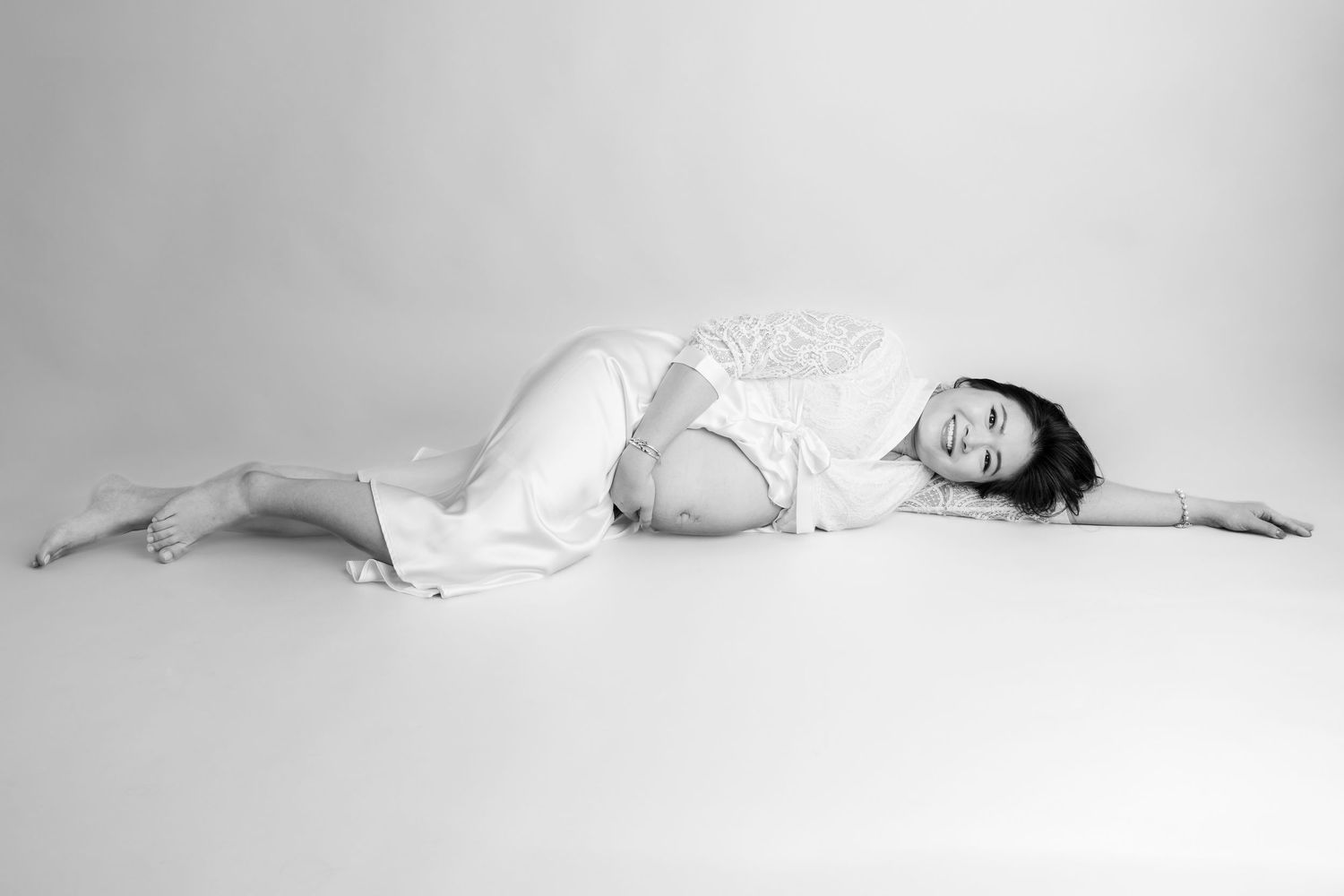 Black and white maternity photo of a pregnant woman lying gracefully on the floor in a white dress.