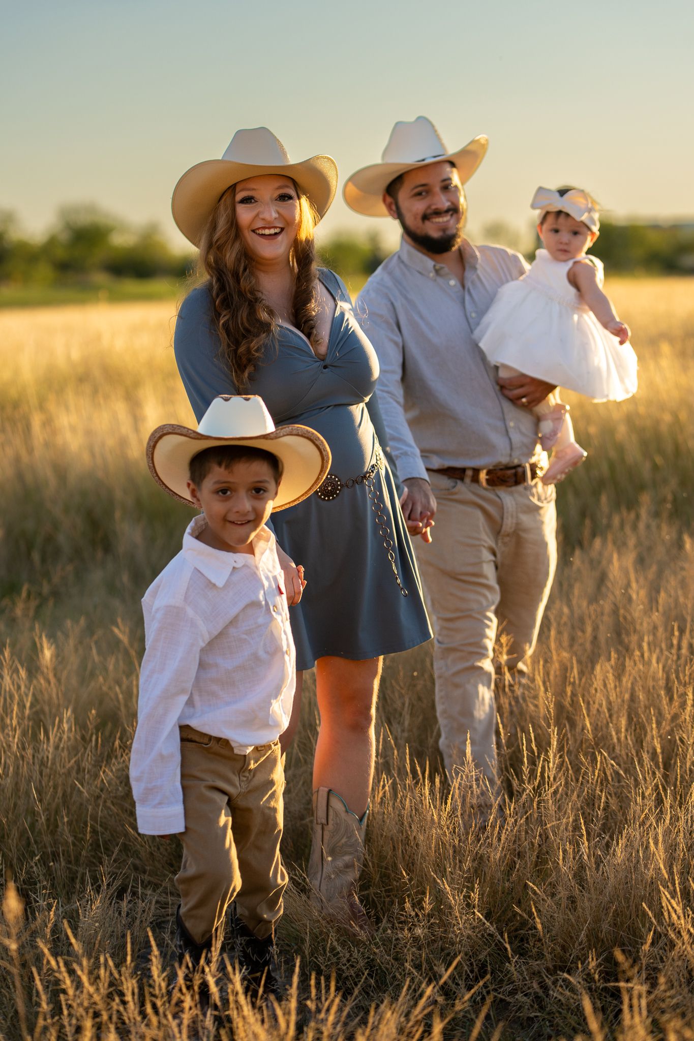 Family photograph of a Texas family with newborn wearing cowboy hats walking together through golden wheat field during sunset golden hour.