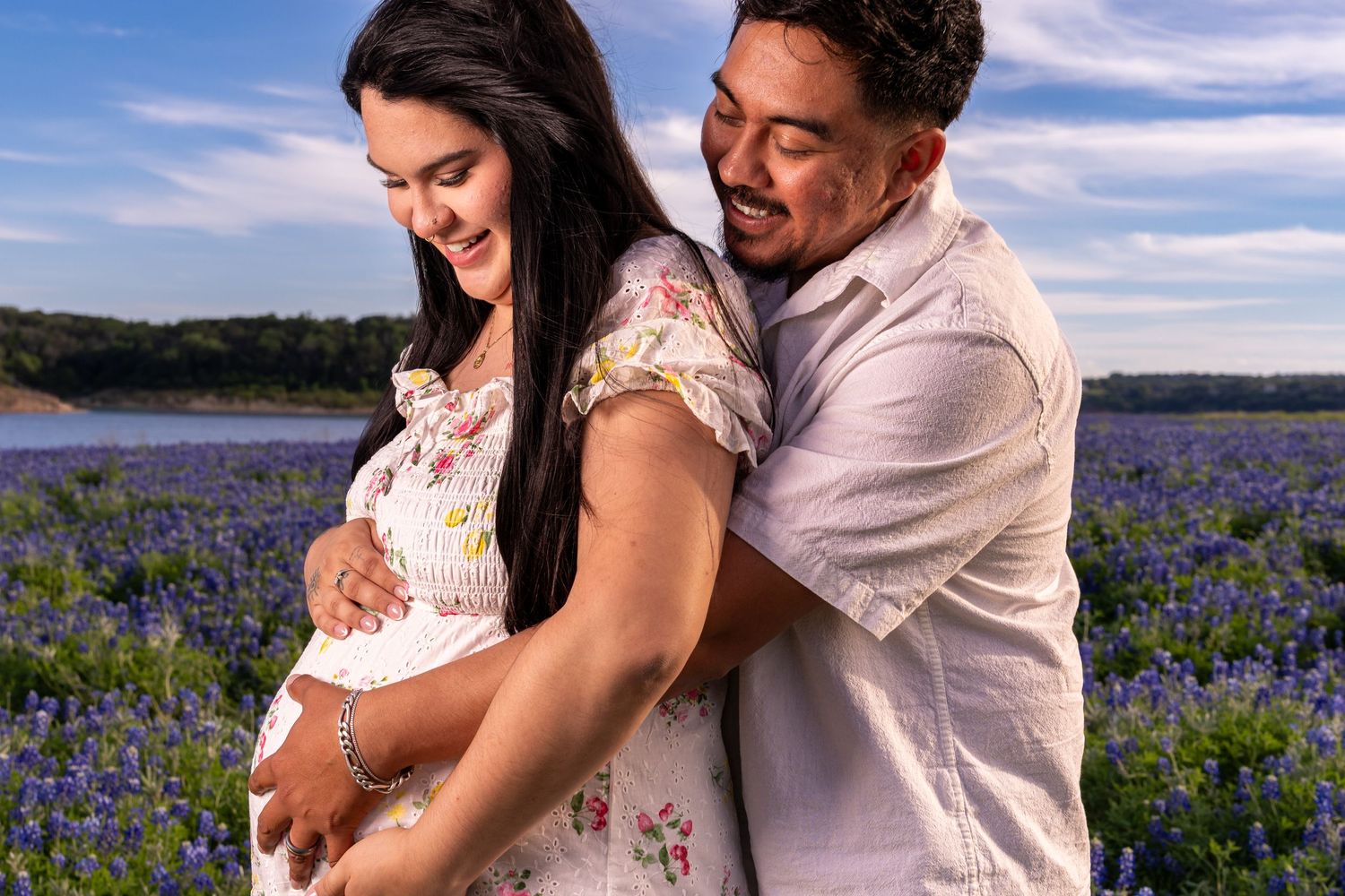 Couple sharing a tender moment in a bluebonnet field, woman cradling pregnant belly under a blue sky.