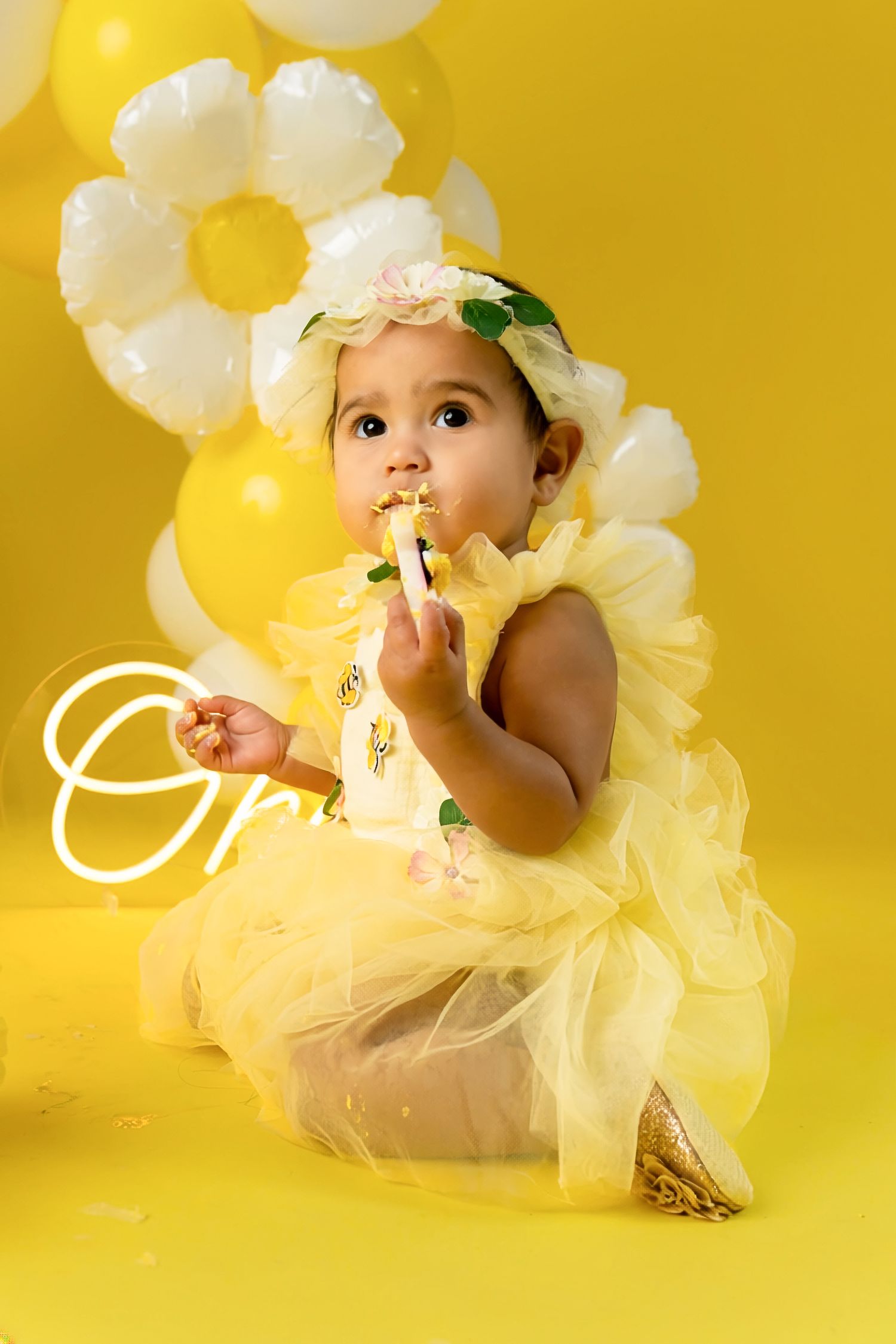 A baby girl in a yellow tutu dress and floral headband sits against a yellow balloon backdrop, eating cake.