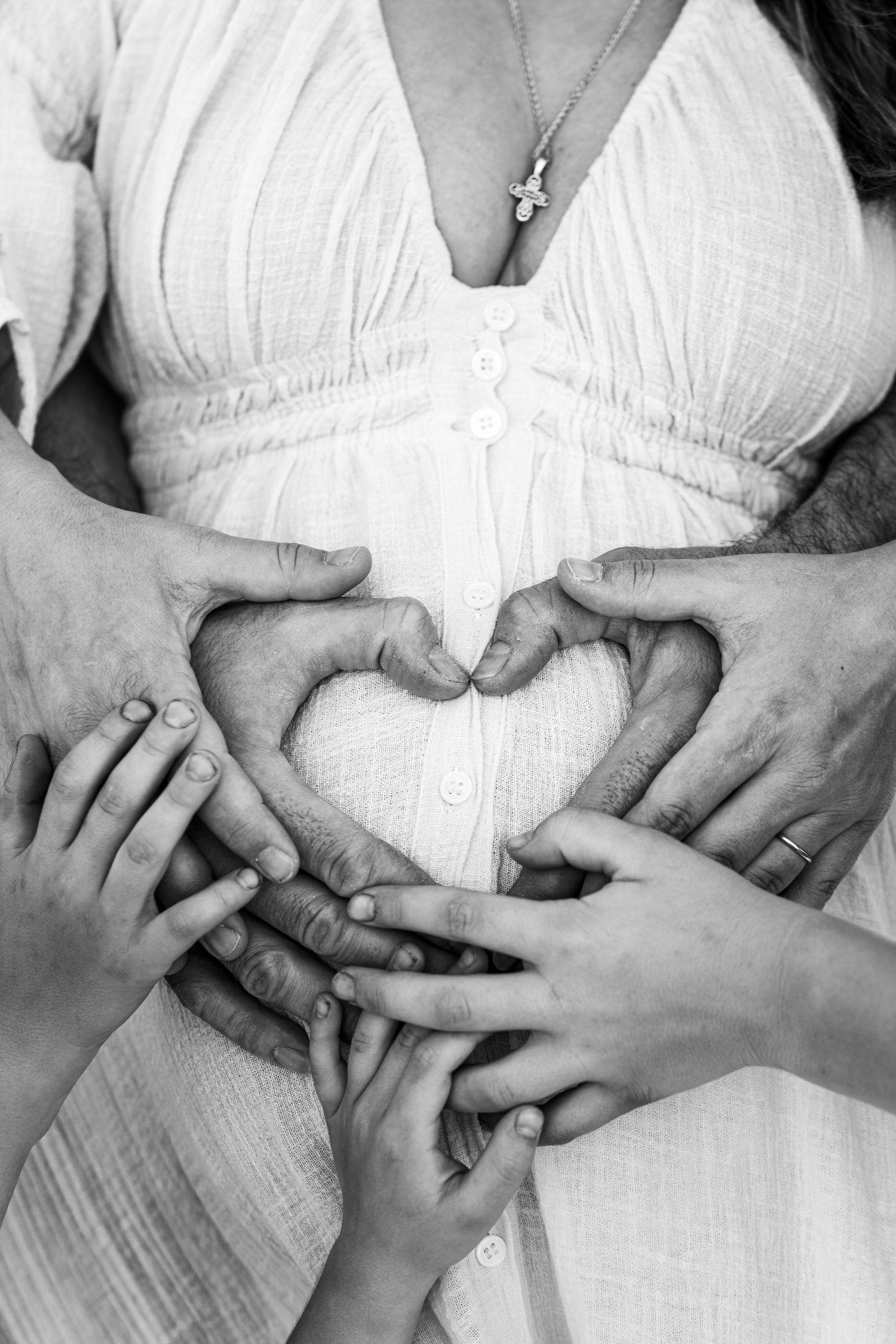 Black and white photo of hands forming a heart shape over a pregnant belly in a white dress.