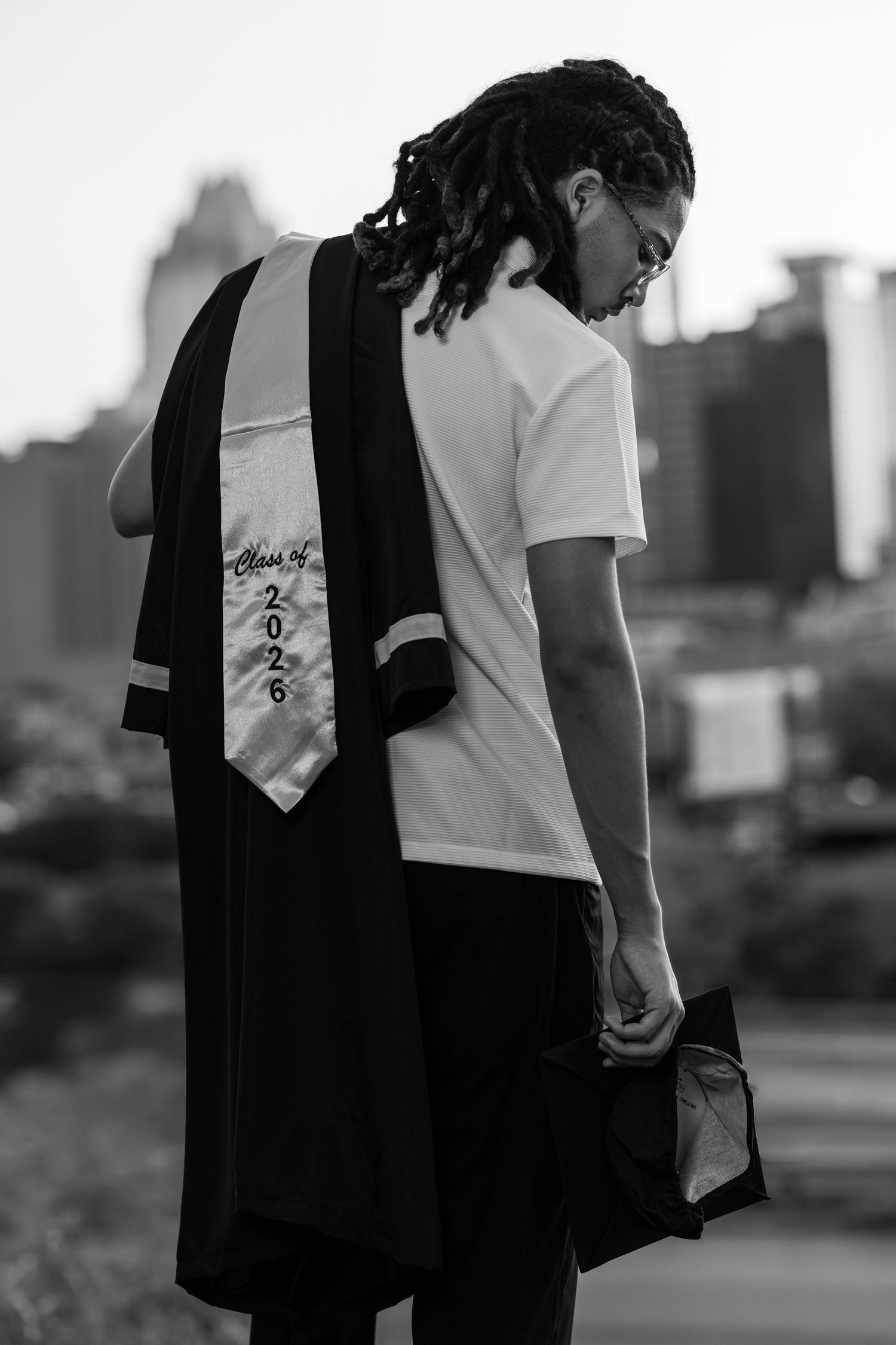 Black and white photo of a graduate with dreadlocks wearing a Class of 2026 stole over a white t-shirt.