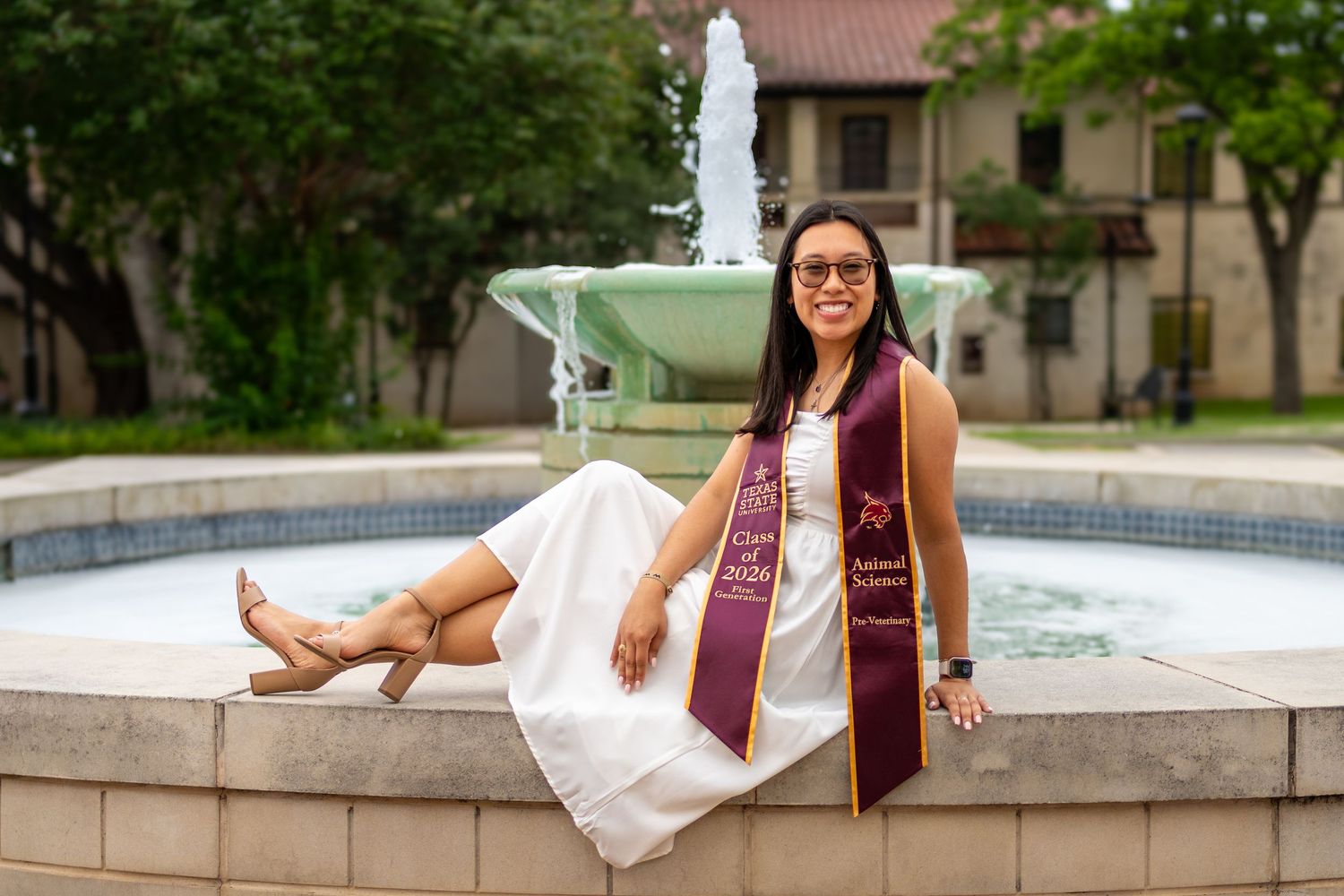 Graduate in white dress and maroon stole poses by fountain on campus for graduation photos.