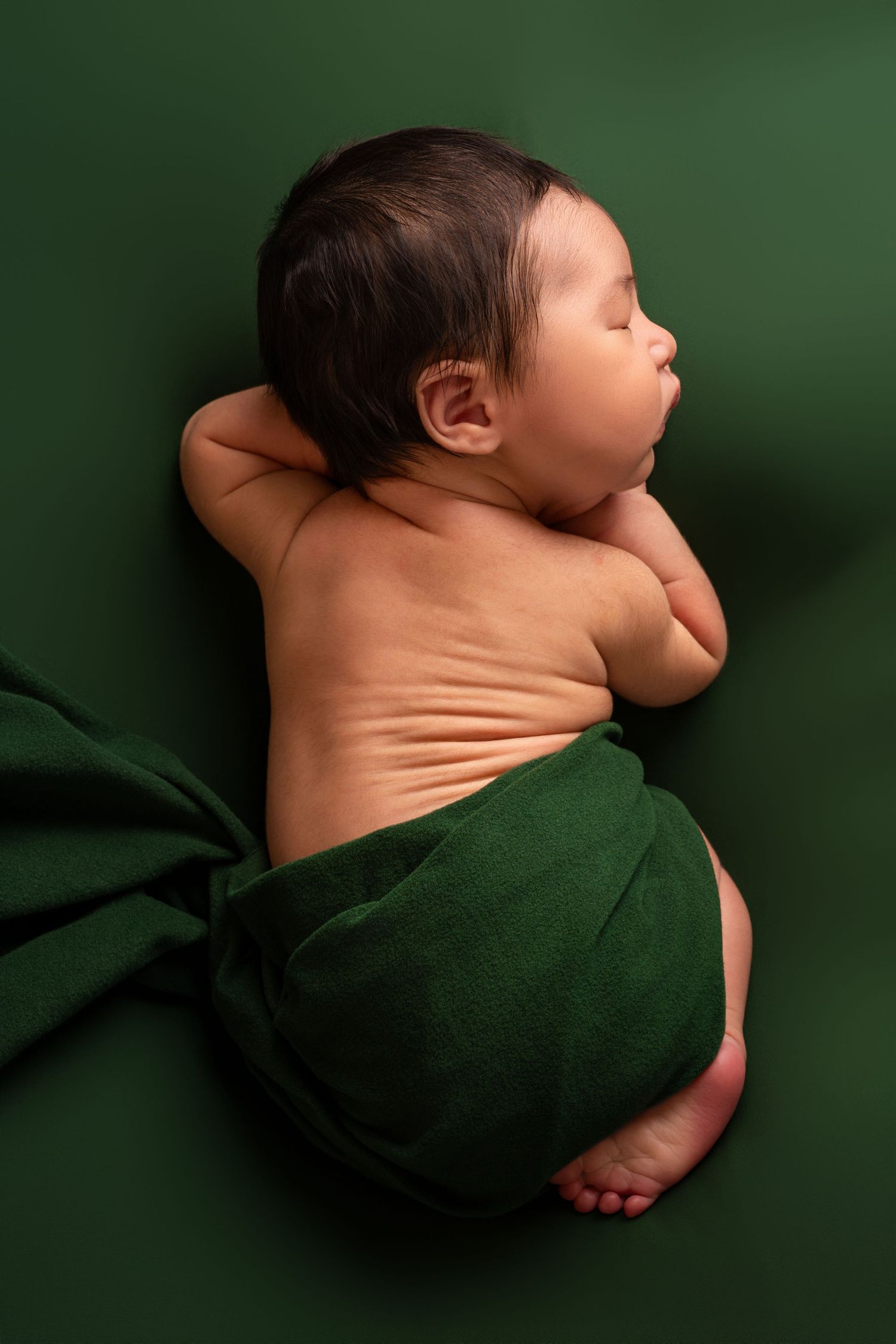 Sleeping newborn baby with dark hair curled on its side, wrapped in a deep green cloth against a matching green backdrop.