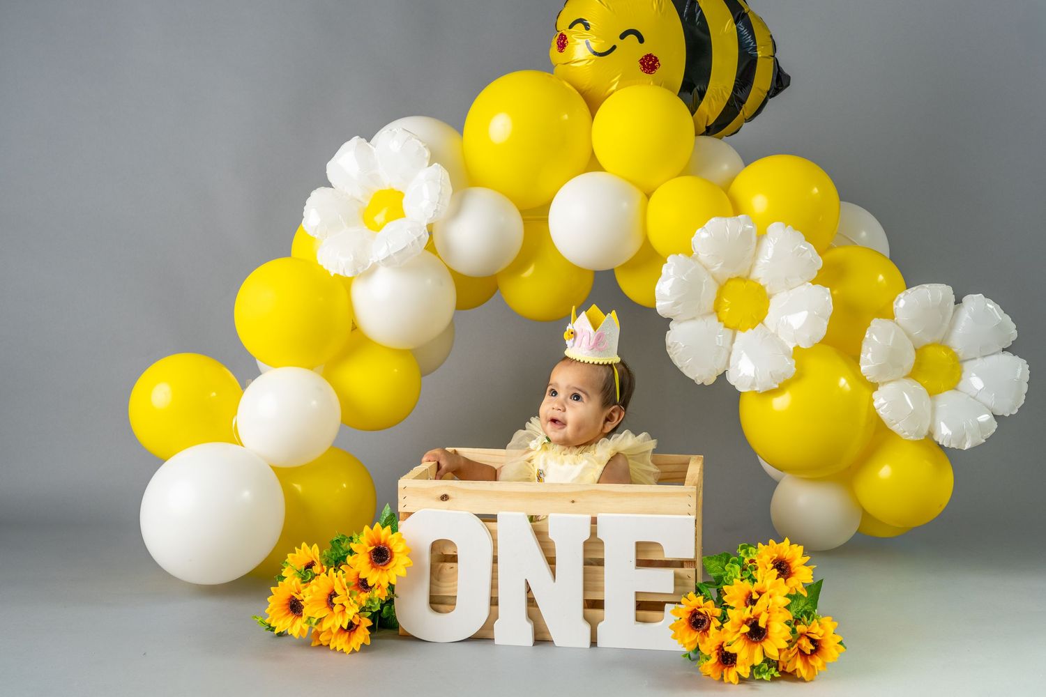 Baby's first birthday cake smash with yellow and white balloon arch, bee balloon, daisy flowers, and ONE sign.
