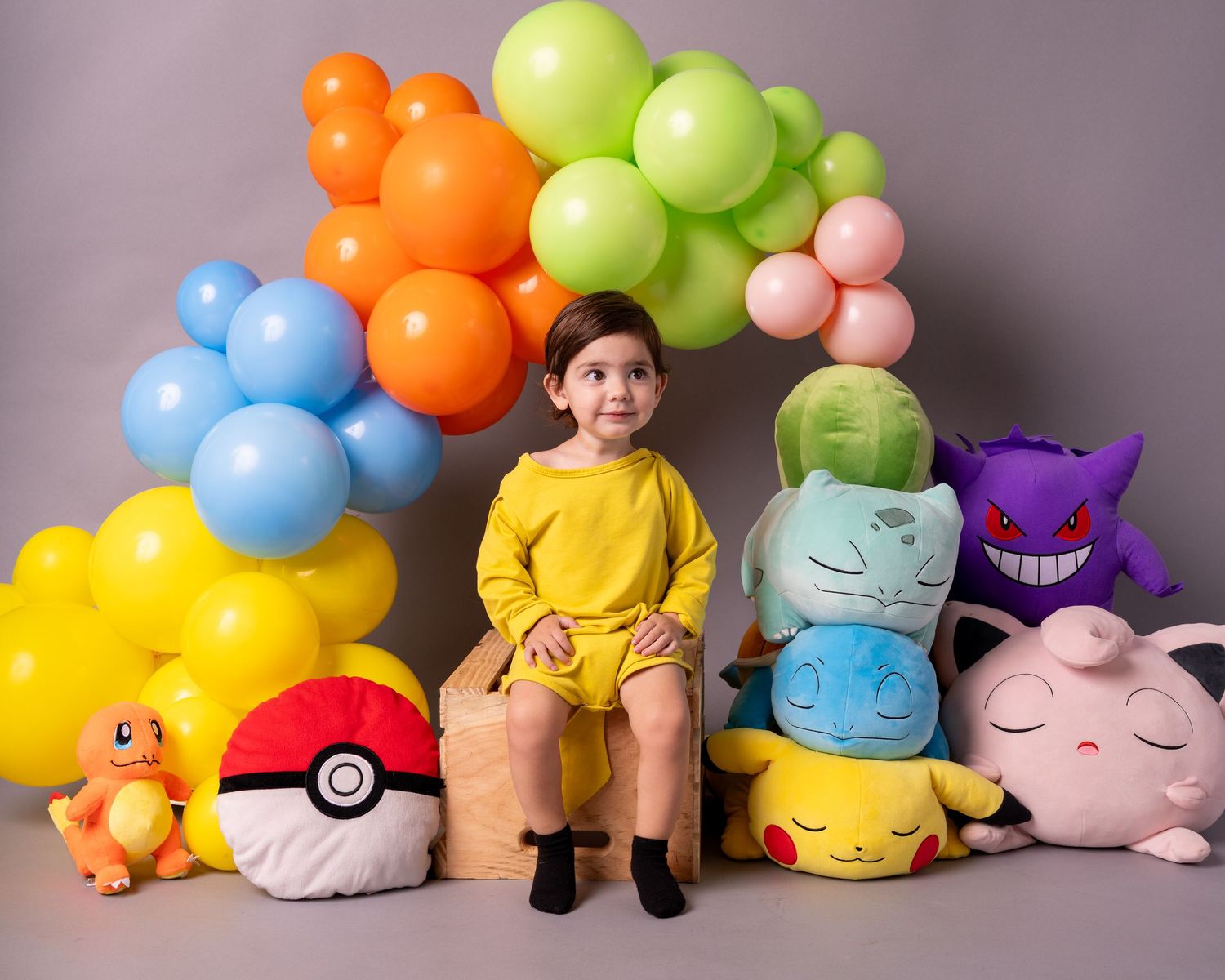Child in yellow outfit poses with Pokemon stuffed animals under a vibrant balloon garland.