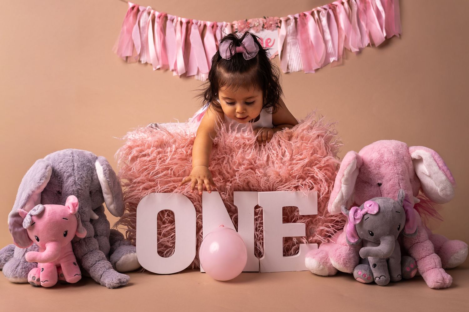 A pink and gray themed baby photo session setup with stuffed elephants and decorative tassels arranged on the floor.