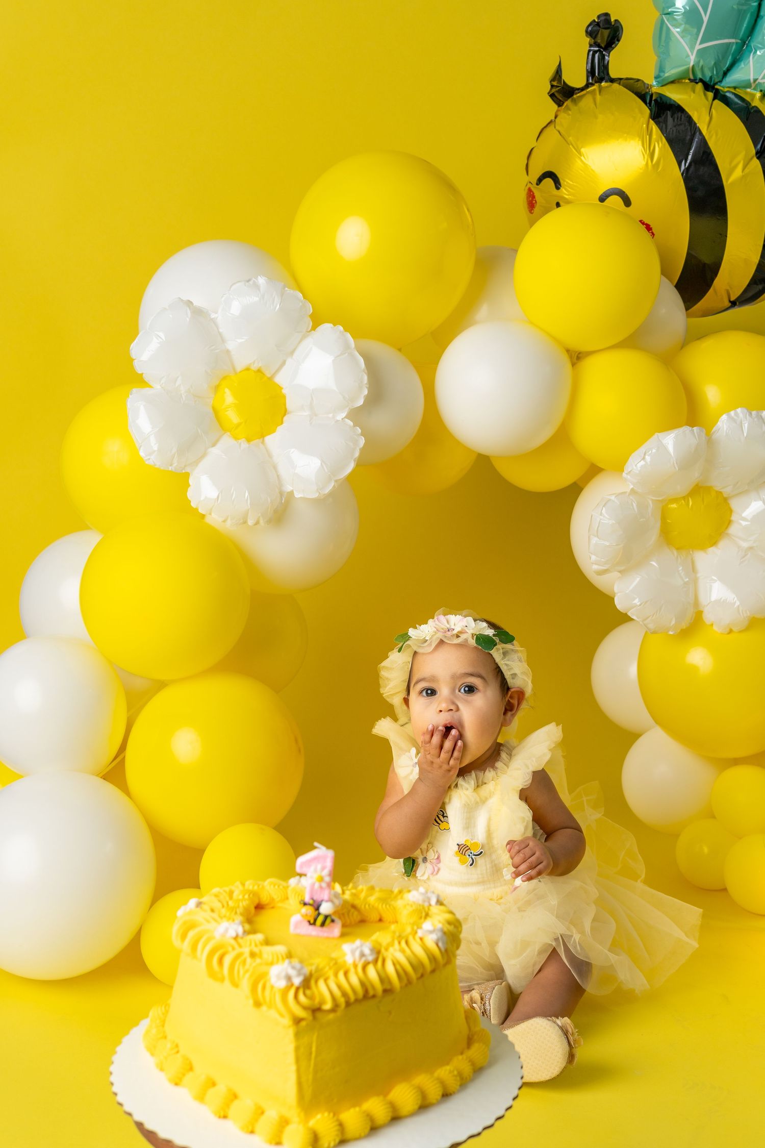 Baby girl in yellow tutu celebrates first birthday with yellow cake, bee balloon, and daisy balloon garland backdrop.