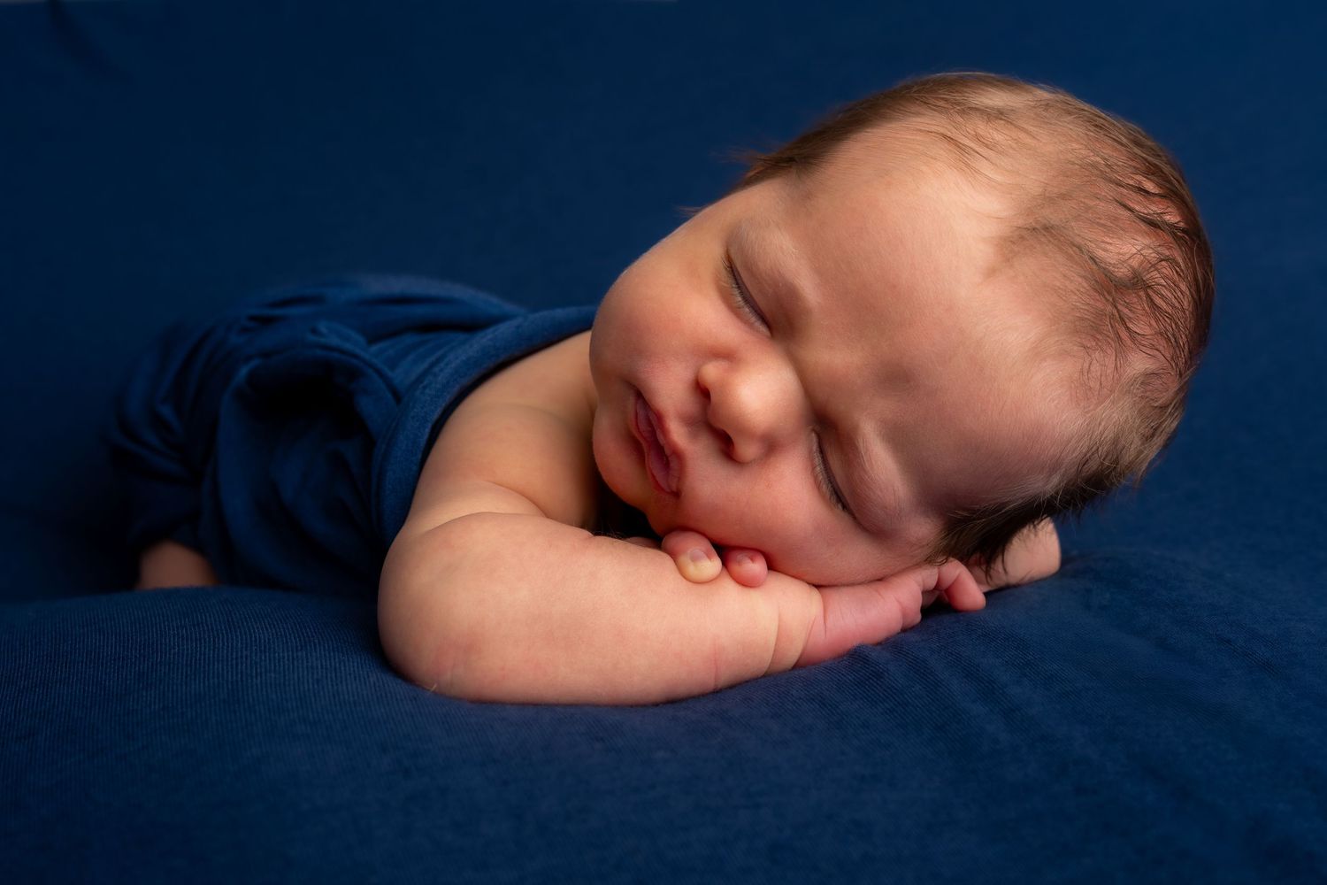 Peaceful newborn baby sleeping on blue fabric, wearing a blue outfit, with tiny hands tucked under cheek.