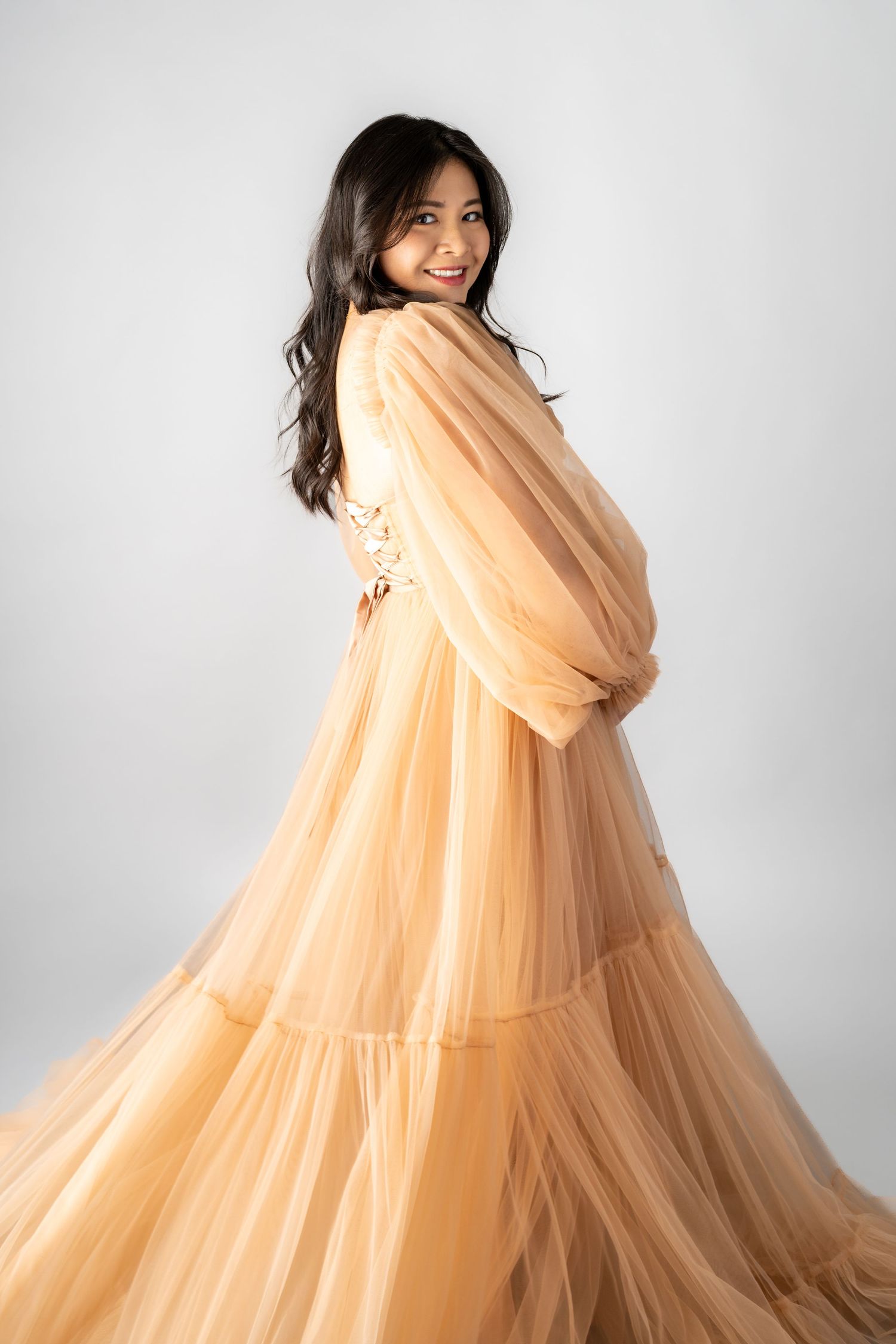 Woman in flowing peach tulle gown twirling gracefully against white background, smiling over shoulder.