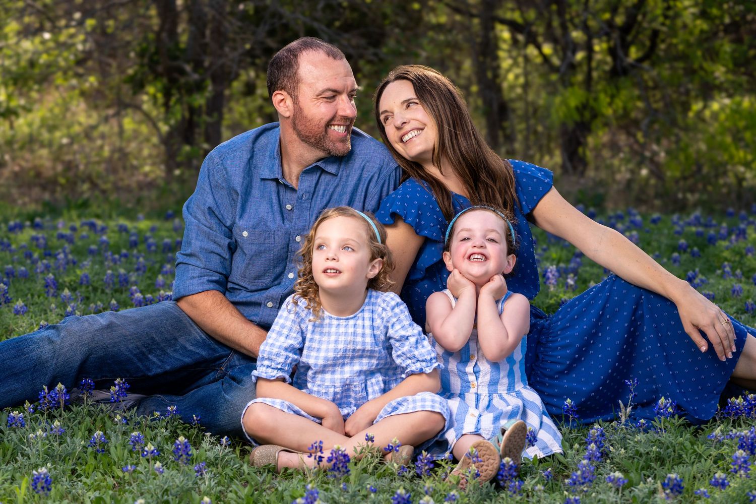 A family wearing coordinating blue outfits sits together on grass surrounded by blooming wildflowers in a natural setting.