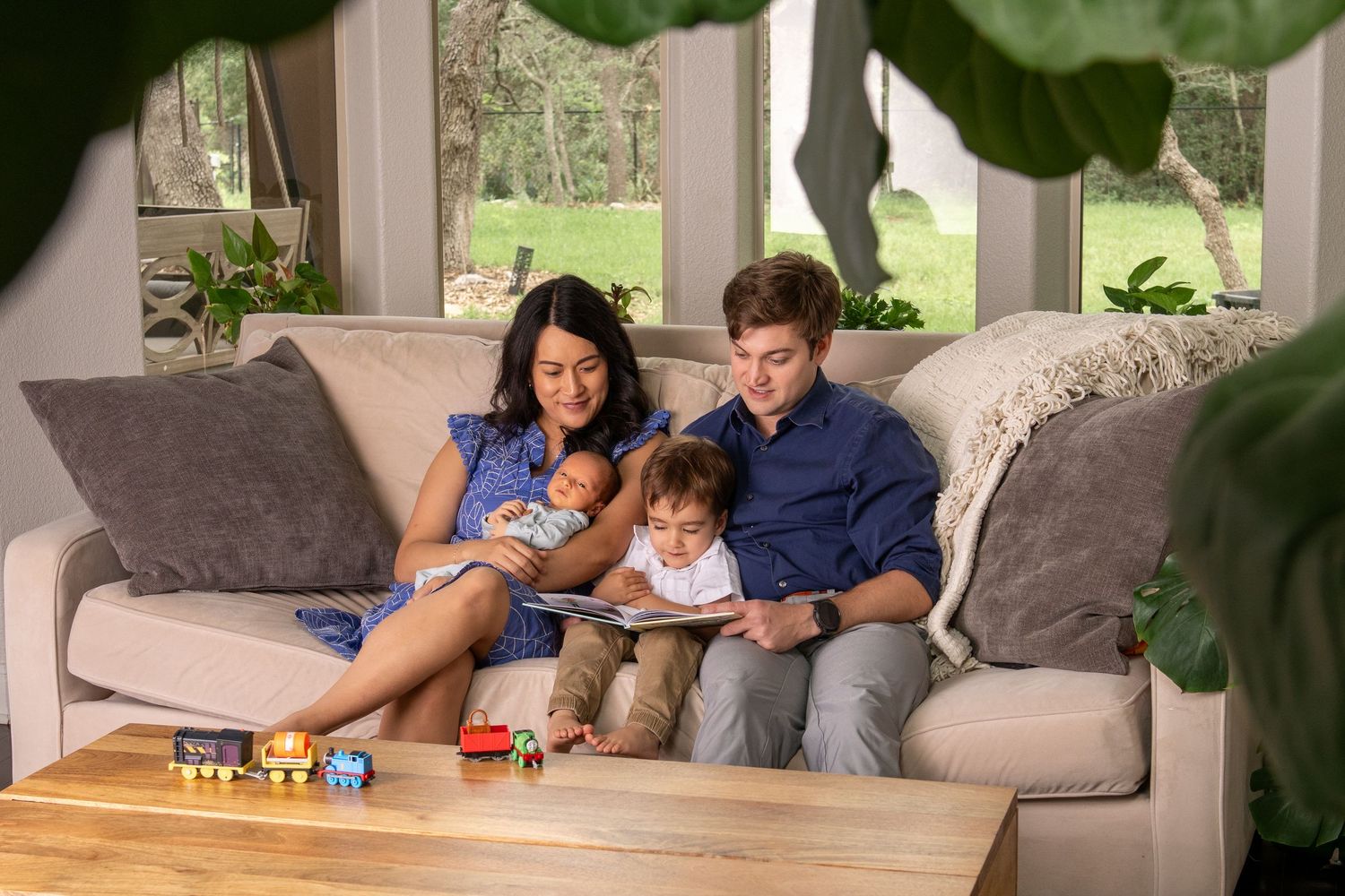 Happy family with newborn and toddler sitting together on a cozy couch reading a book in a bright living room.