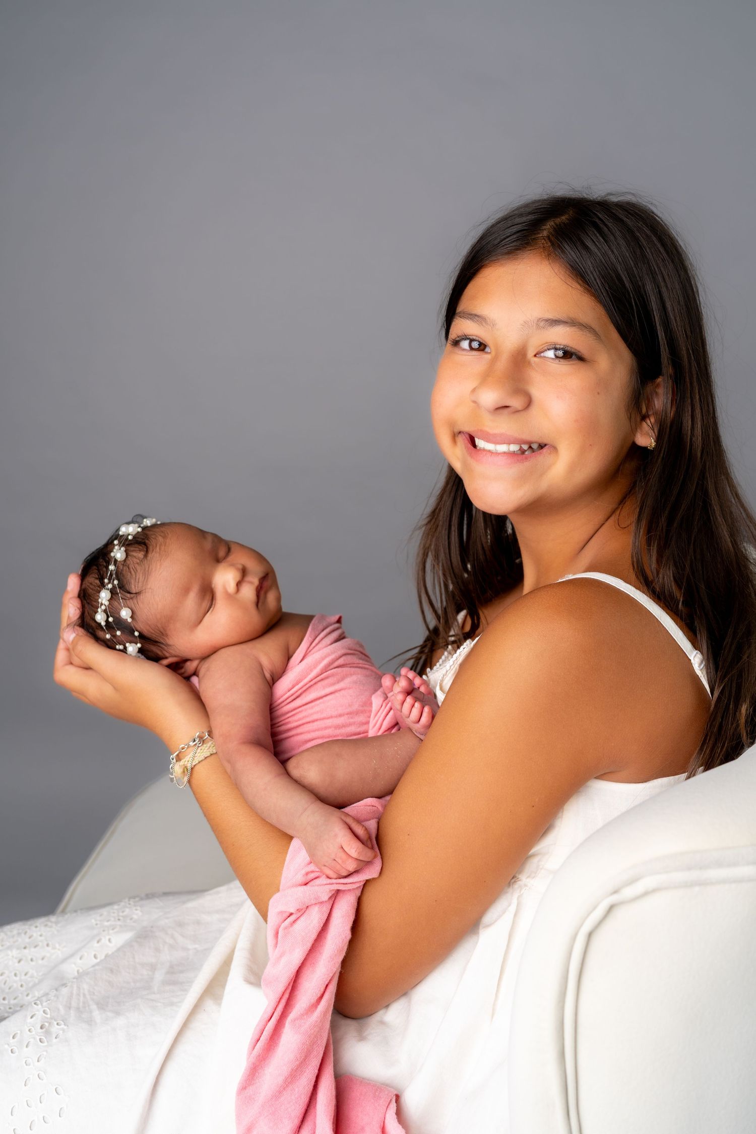 Smiling young woman in white dress holding a sleeping newborn baby wrapped in pink with a floral headband.
