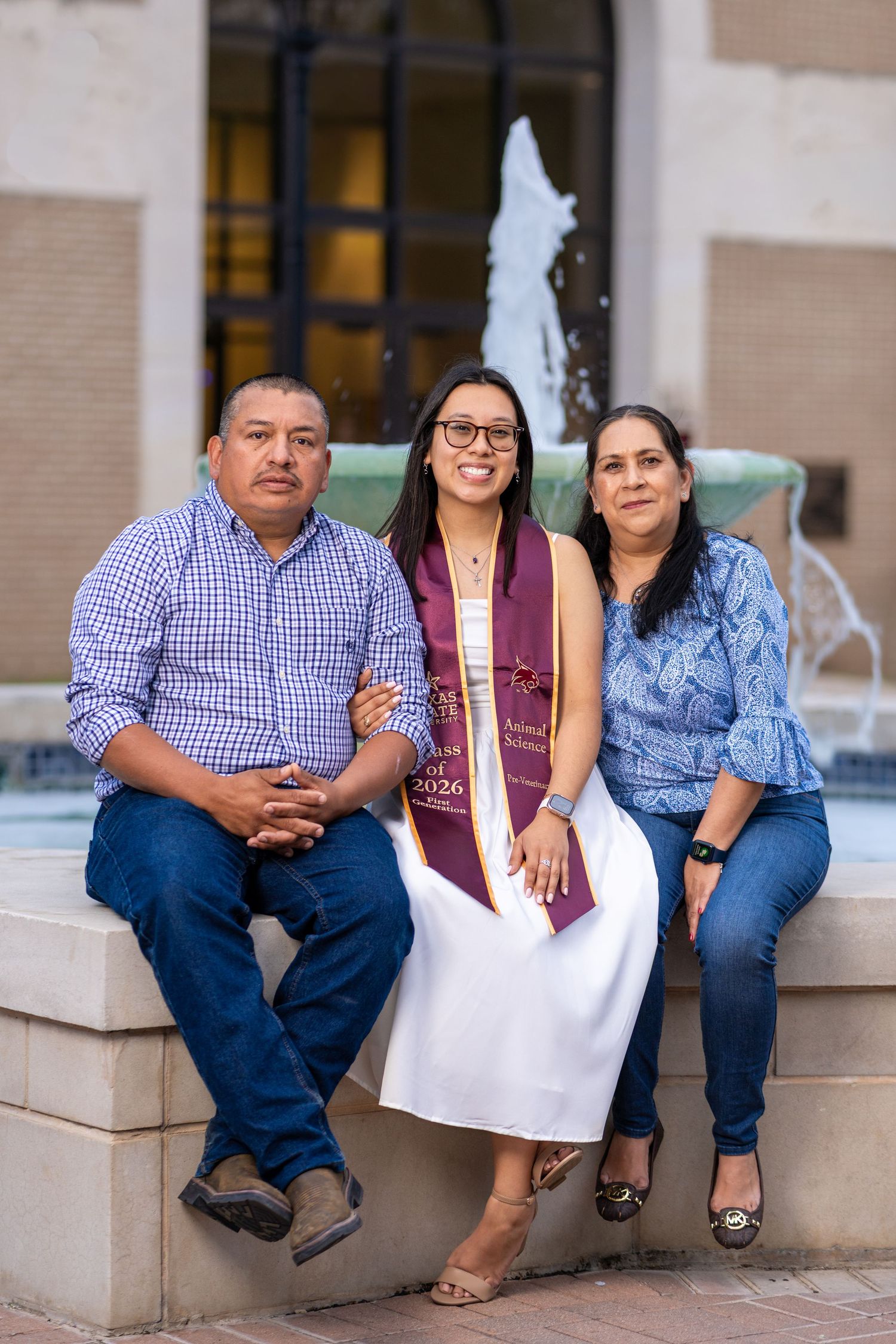 A graduate in white dress and purple stole poses with her proud parents by a fountain.
