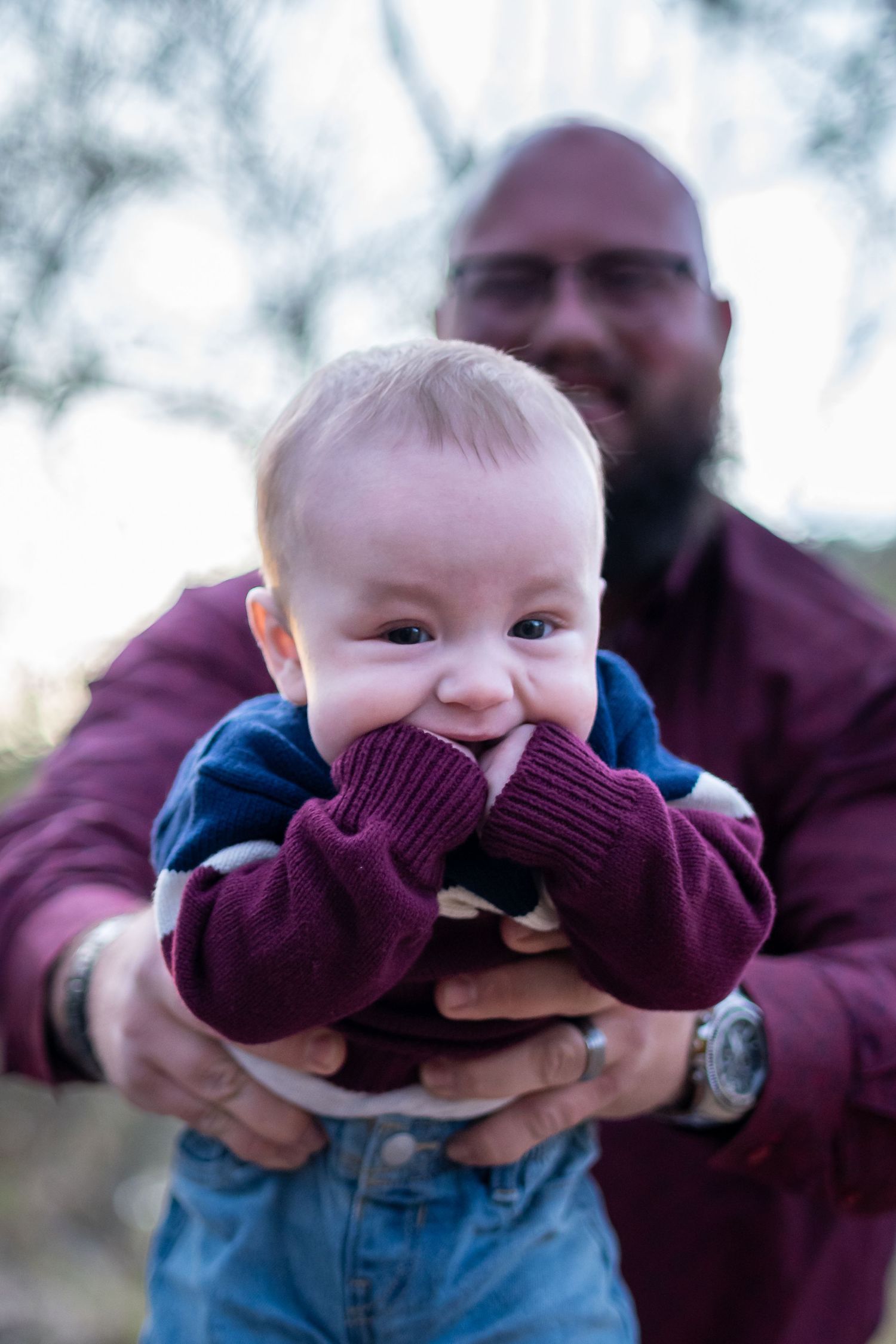 Playful baby wearing burgundy sweater makes silly faces during outdoor family photoshoot.