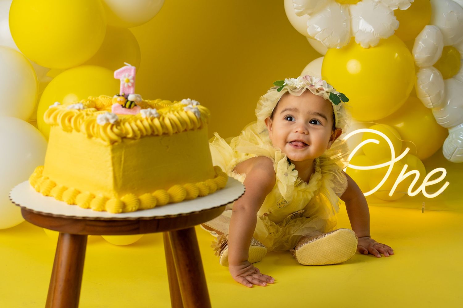 Baby girl in yellow dress celebrates first birthday with yellow cake and balloon backdrop.
