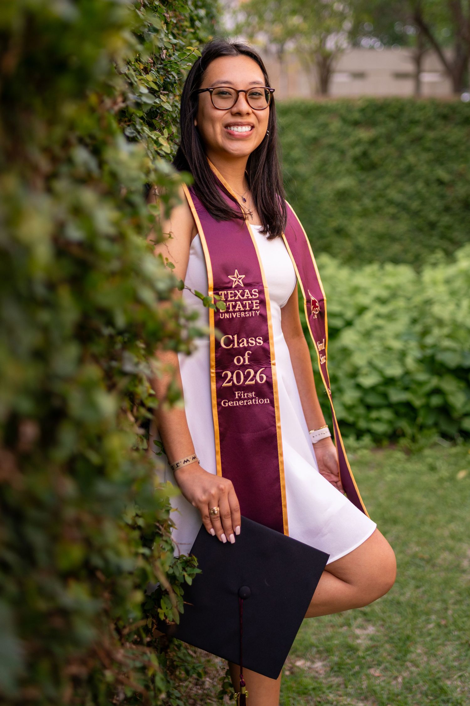 Smiling Texas State University Class of 2026 First Generation graduate wearing maroon sash and white dress outdoors.
