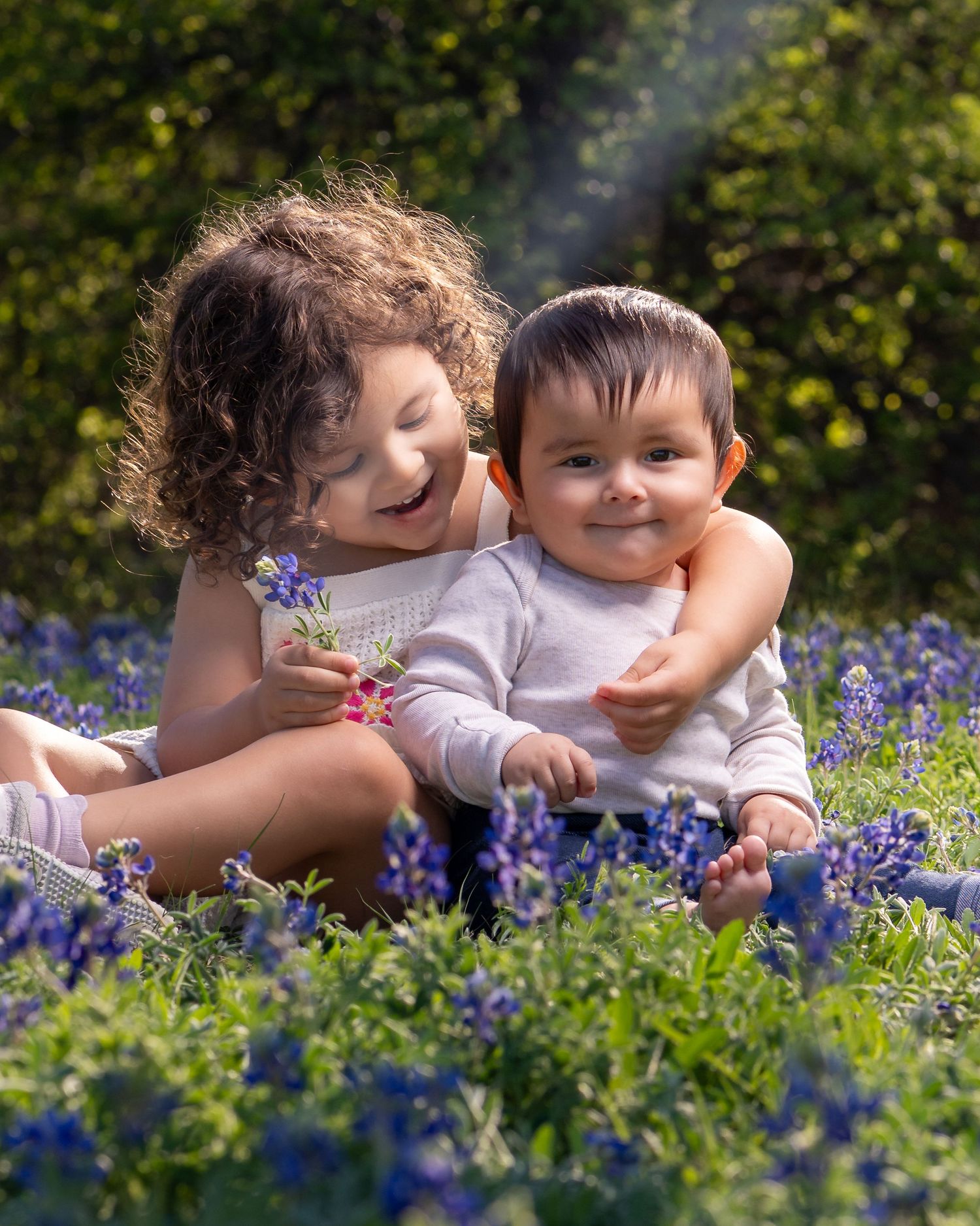 Two small children sitting together in a field of purple bluebonnets and wildflowers during springtime.