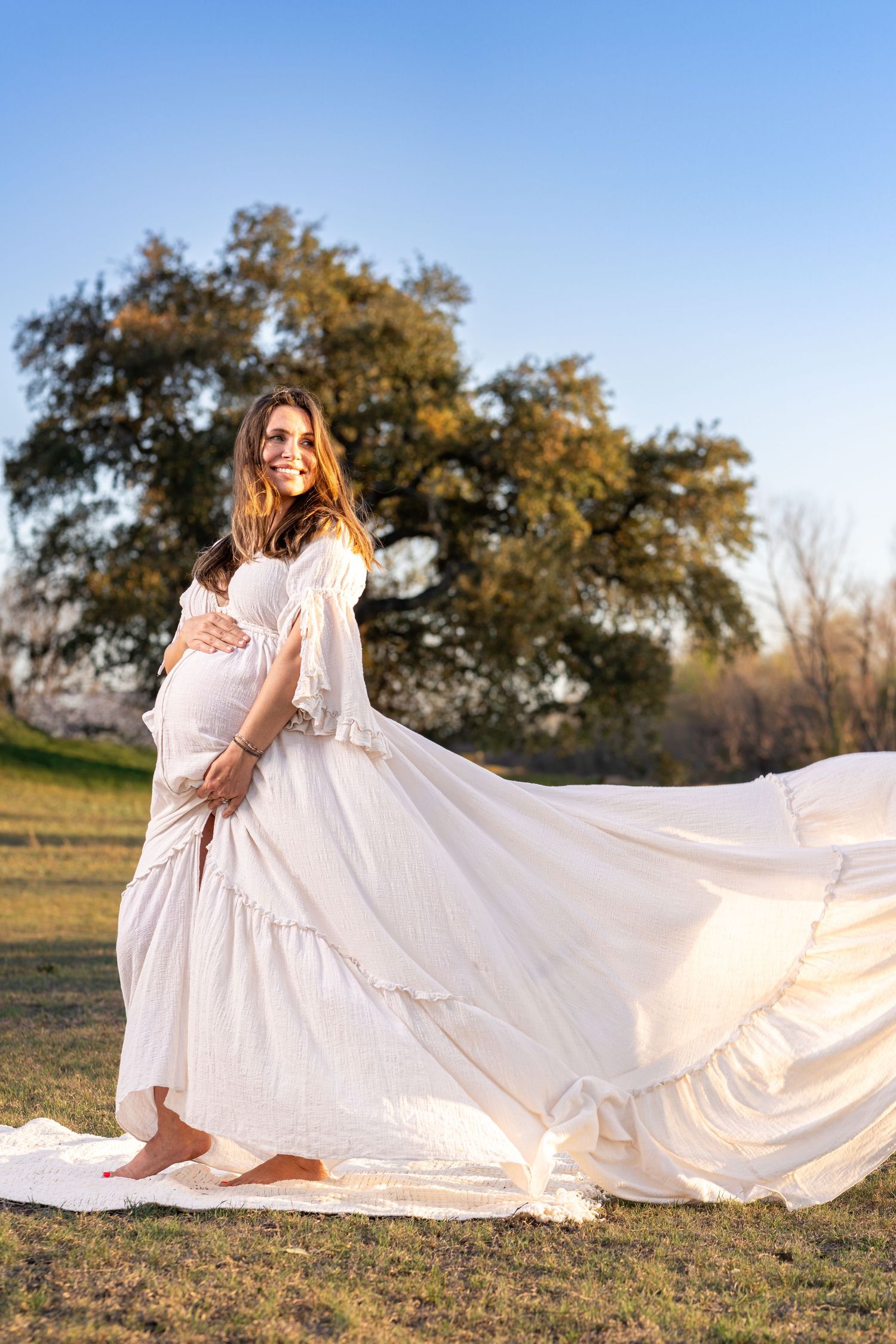 A flowing white maternity dress billows in the breeze during a golden hour outdoor photoshoot with trees in the background.