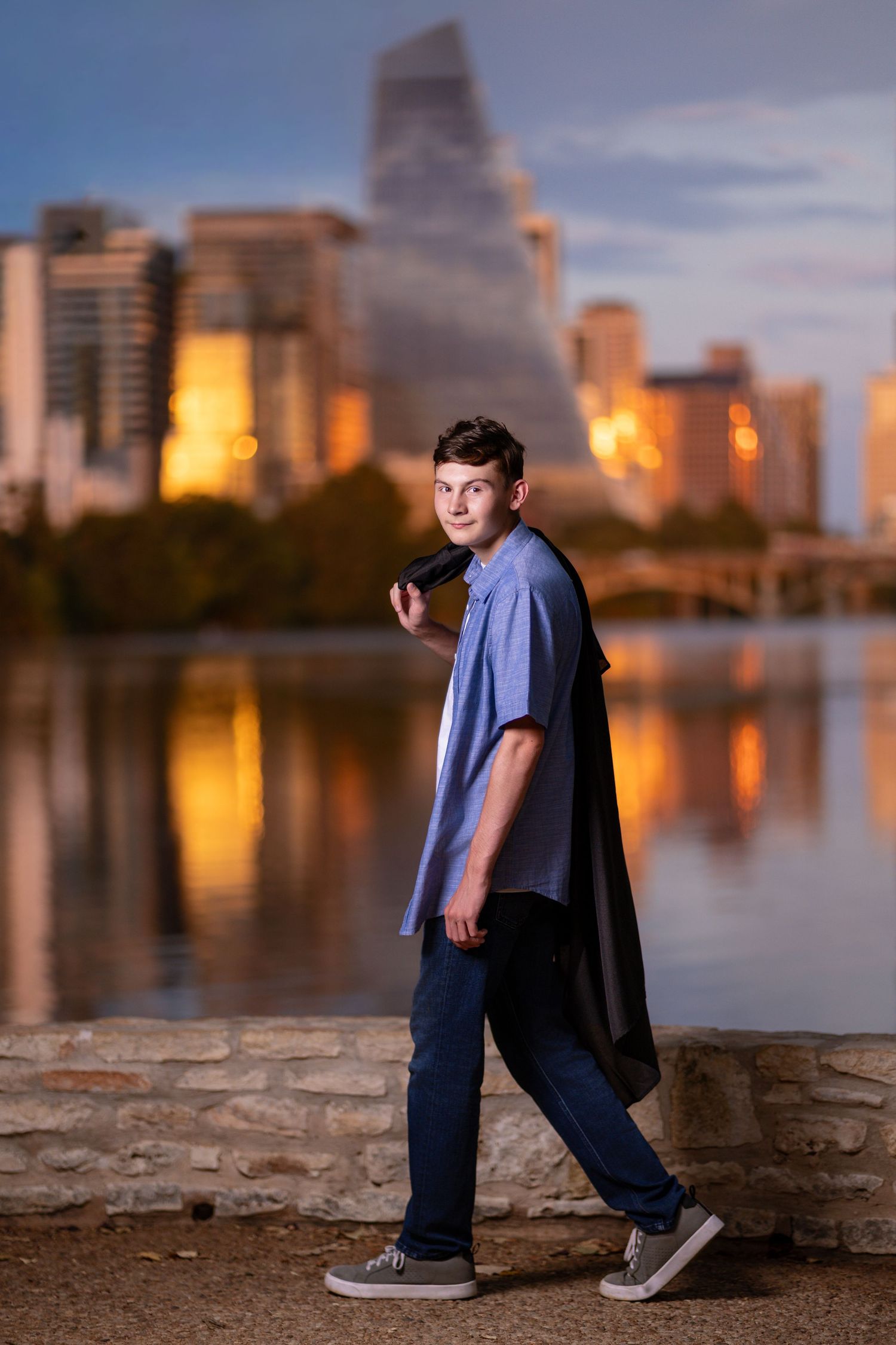 Young man in blue shirt poses by urban waterfront at dusk with city skyline reflections behind him.